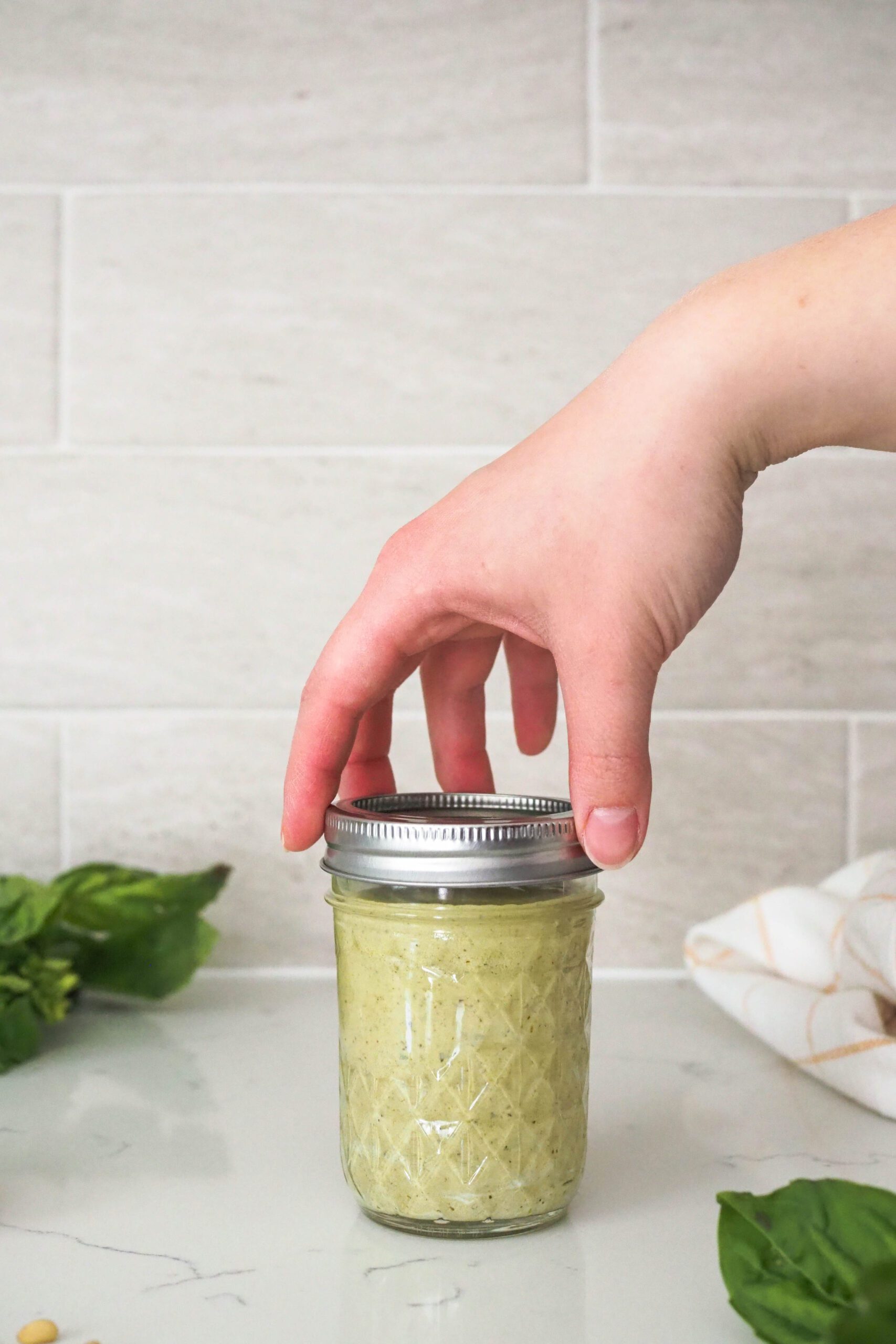 A hand screws on the lid of a jar containing creamy basil pesto.