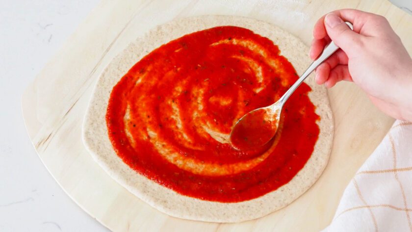 A spoon spreads pizza sauce across a pizza dough round.