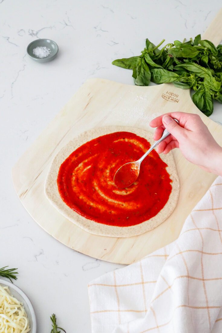 A hand spreads pizza sauce onto a round of pizza dough.