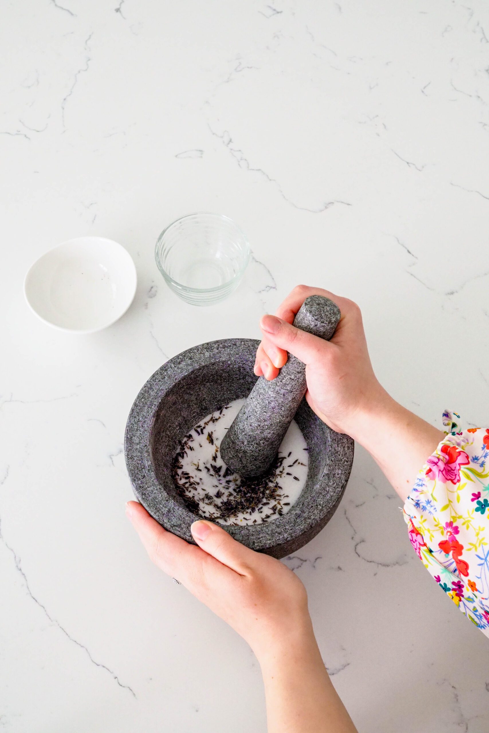 A hand grinds a pestle into a mortar with lavender and sugar.