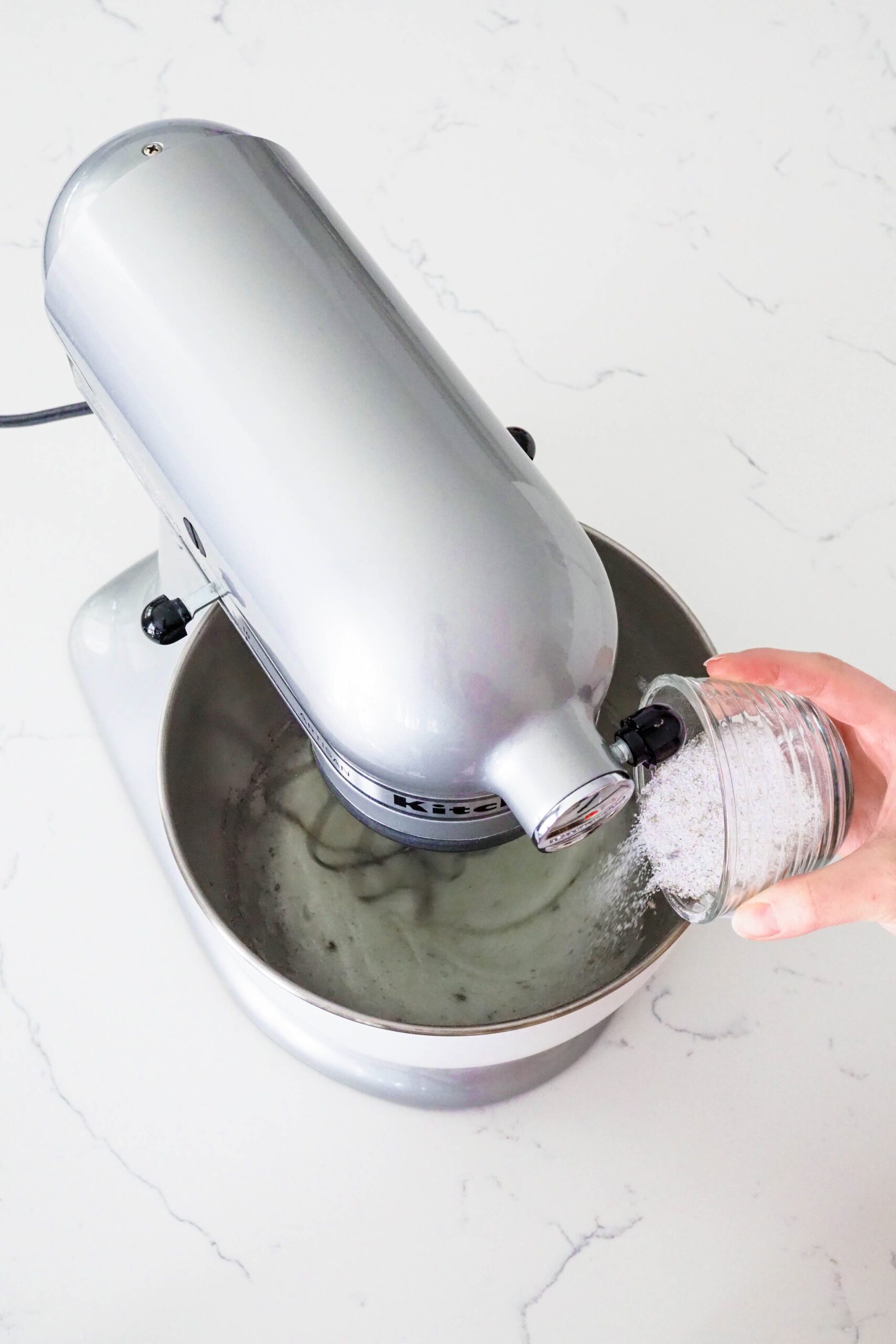 A hand pours lavender sugar into a stand mixer bowl whipping egg whites.
