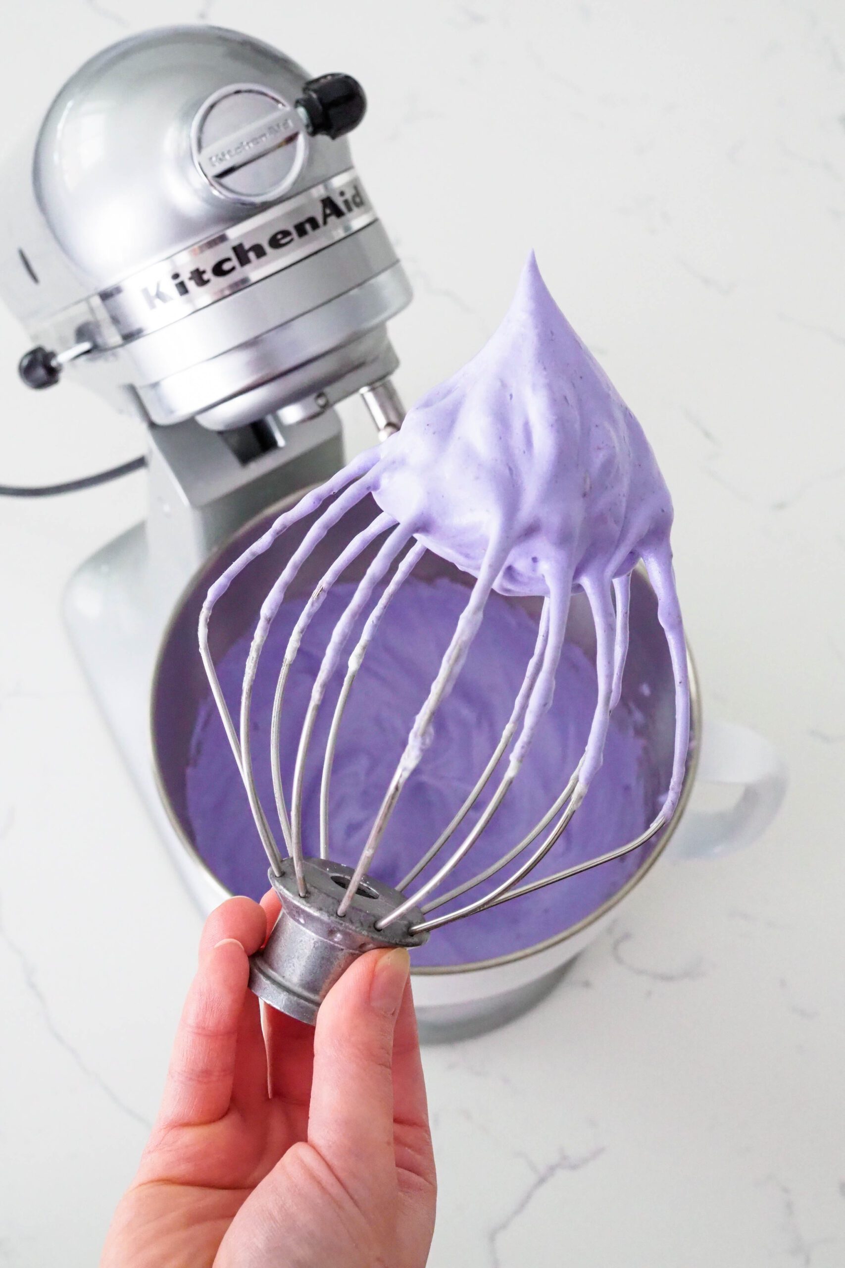 A hand holds a KitchenAid whisk attachment with purple lavender meringue sticking straight up from it.