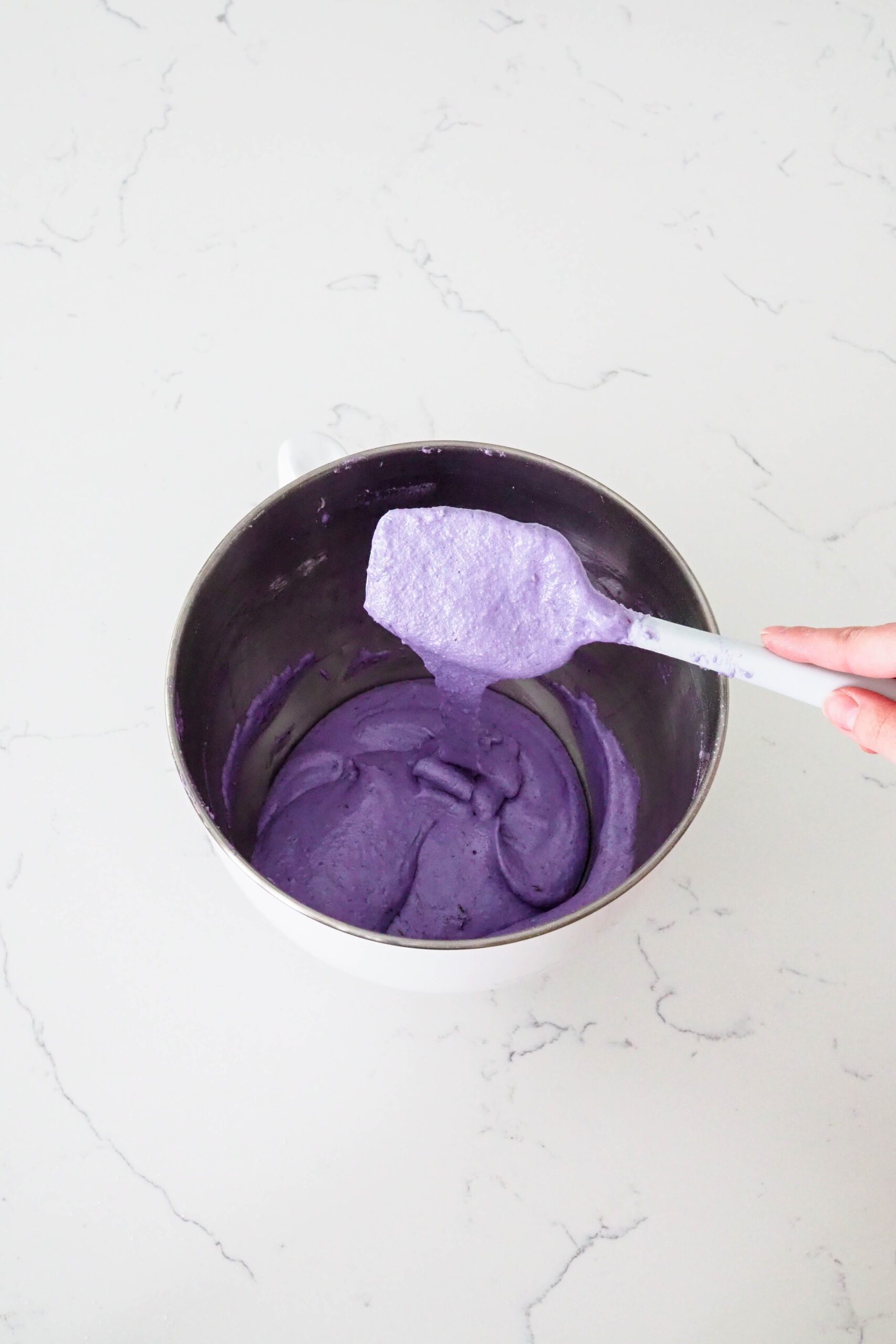 A spatula drips purple macaron batter back into the bowl.