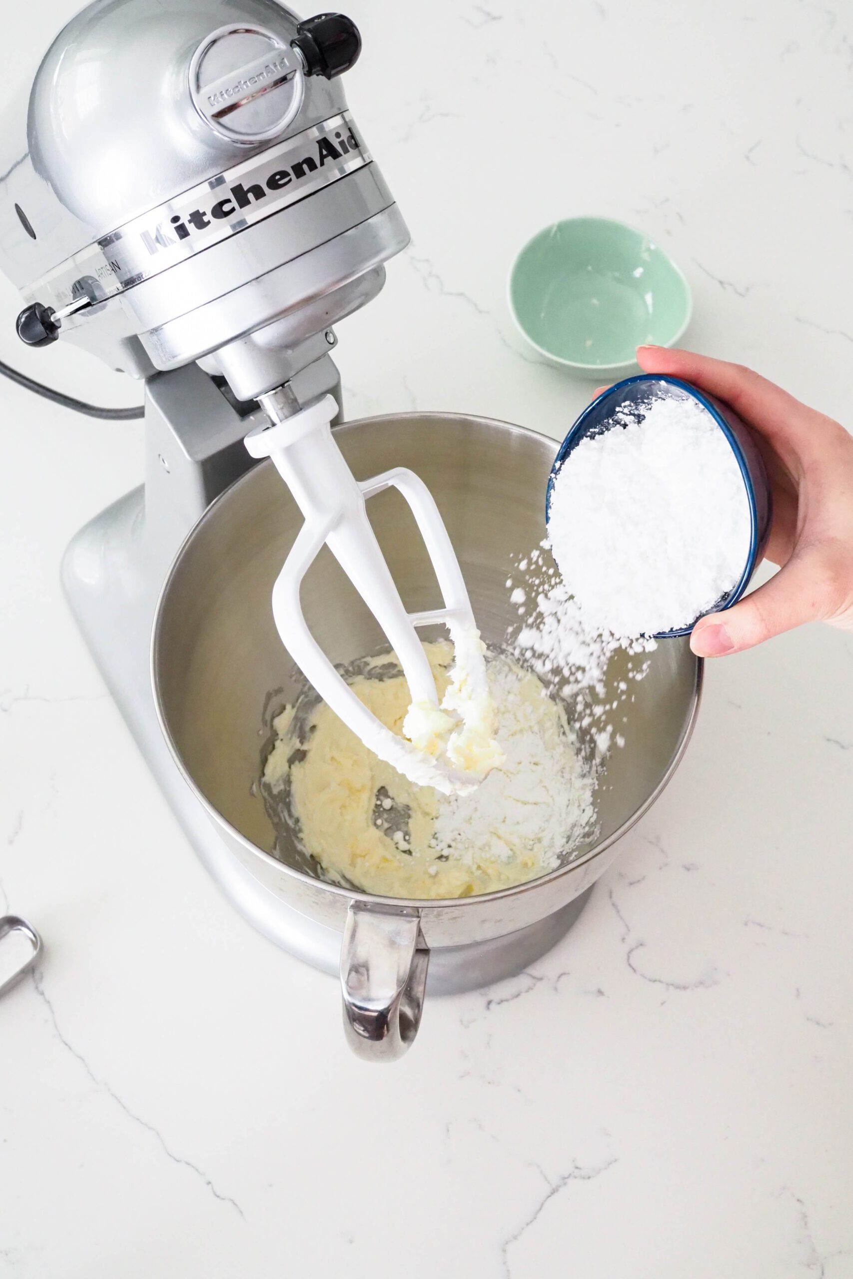 A hand sprinkles powdered sugar into a stand mixer with butter.