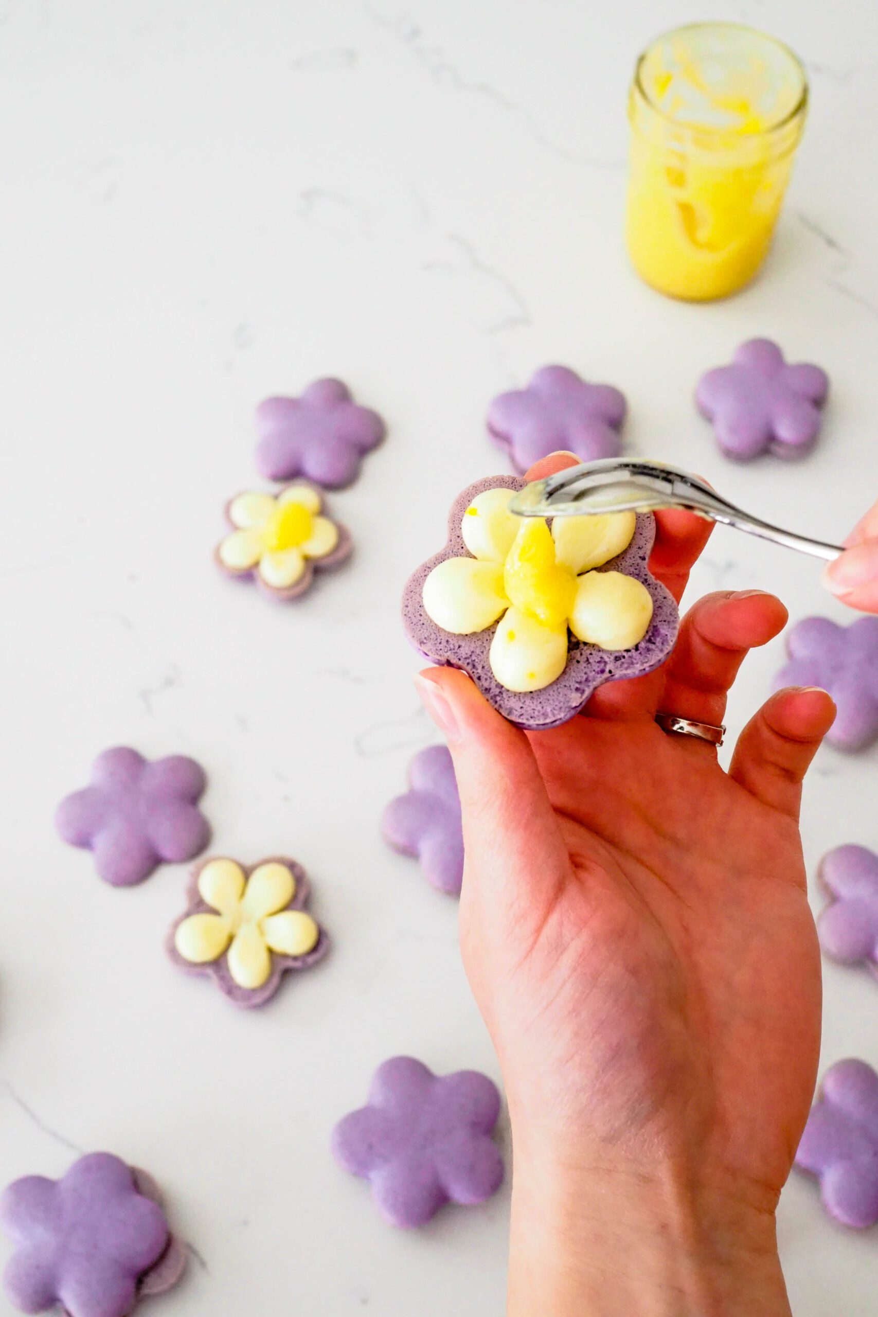 A small spoon adds a dollop of lemon curd to the center of a flower-shaped lemon lavender macaron.