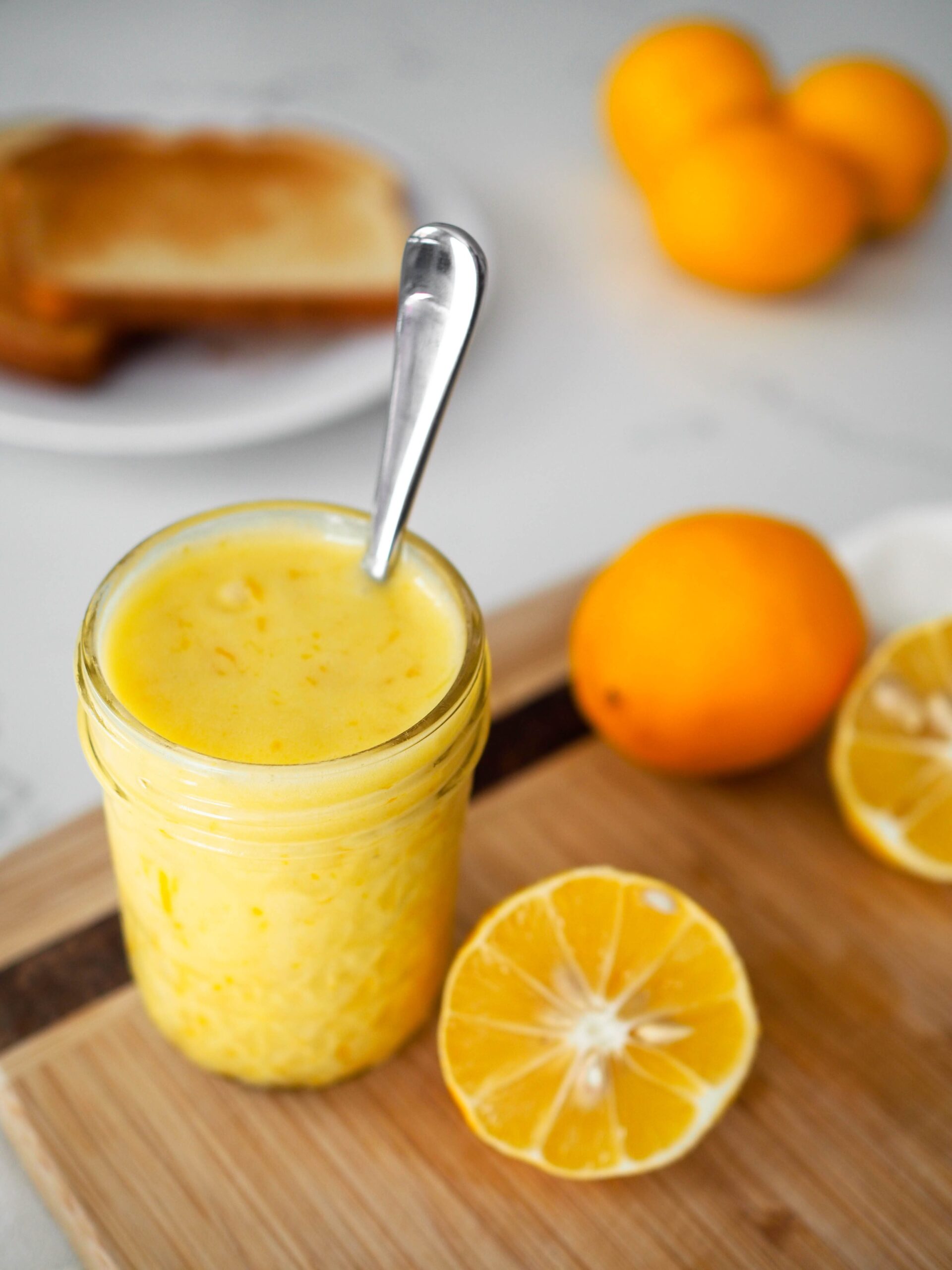 A spoon sticks out of a jar of lemon curd on a wooden cutting board.