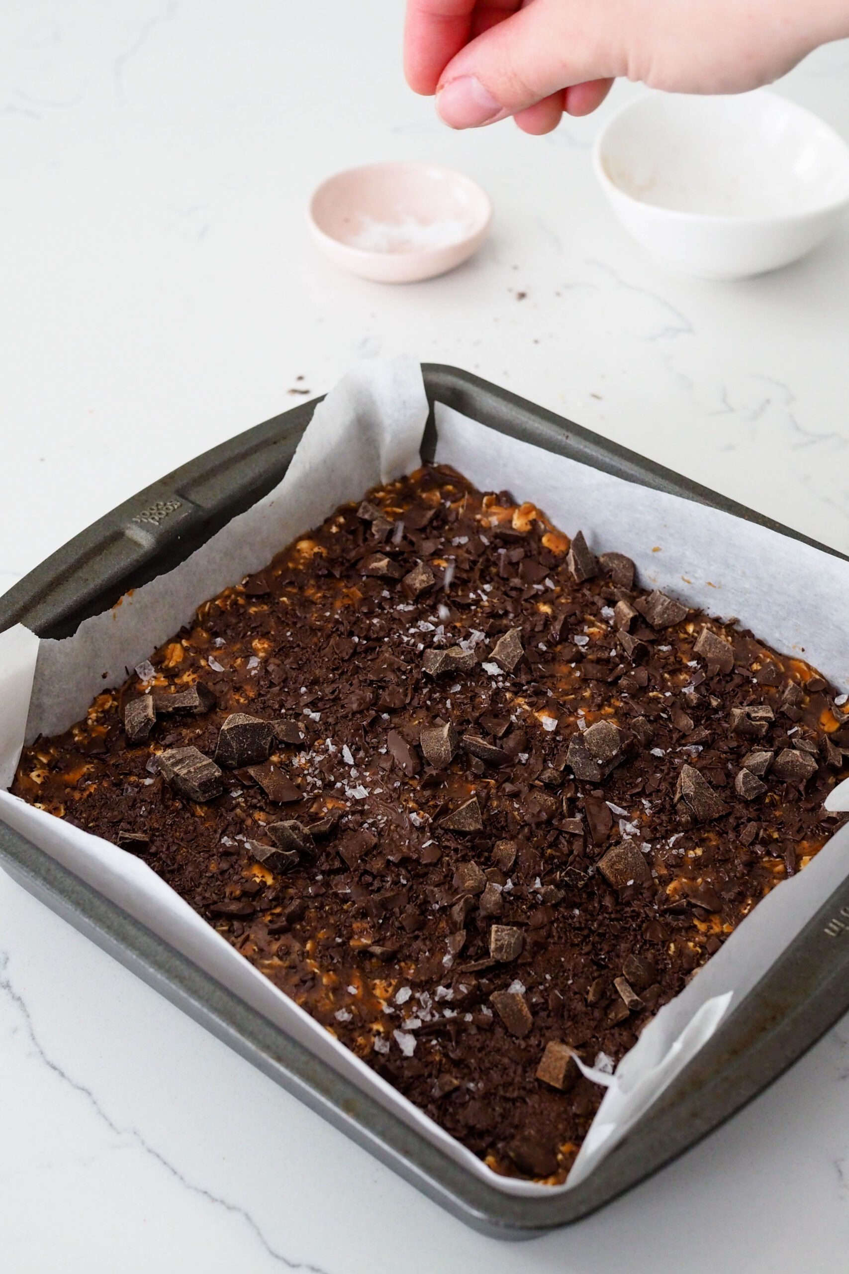 A handle hovers over a pan of homemade protein bars, sprinkling flaky salt onto them.