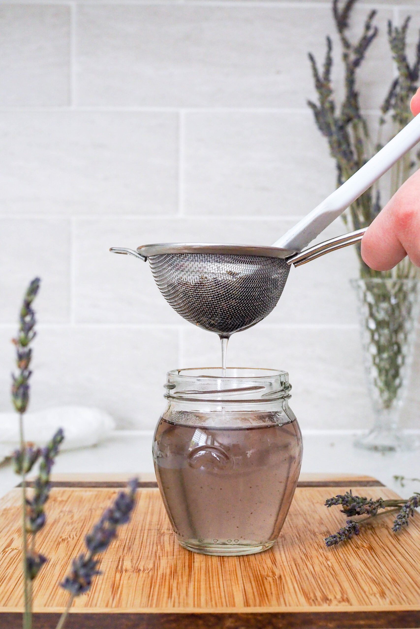 A spatula presses syrup out of lavender buds in a strainer over a jar.