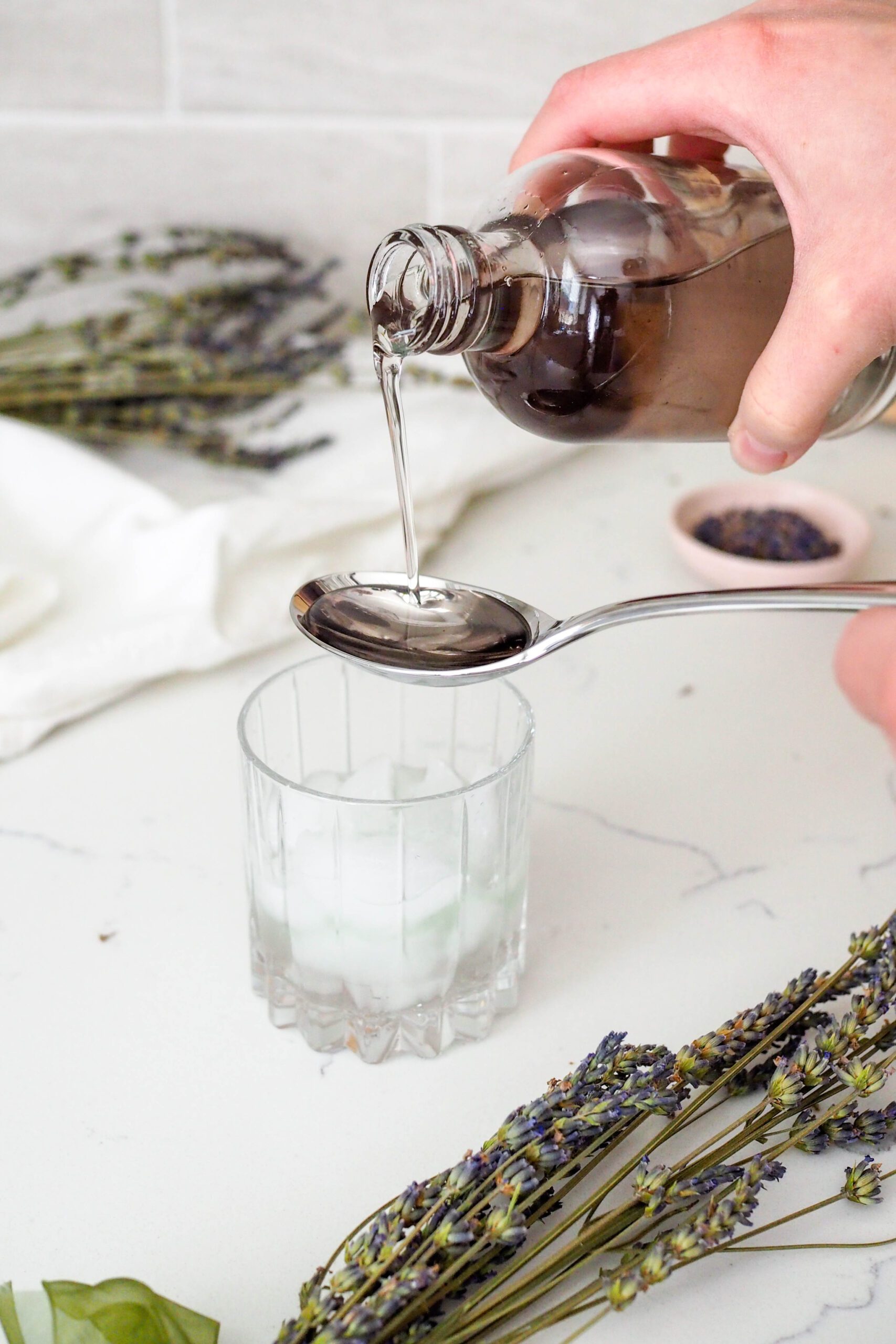 Lavender syrup is poured into a tablespoon over a whiskey glass.
