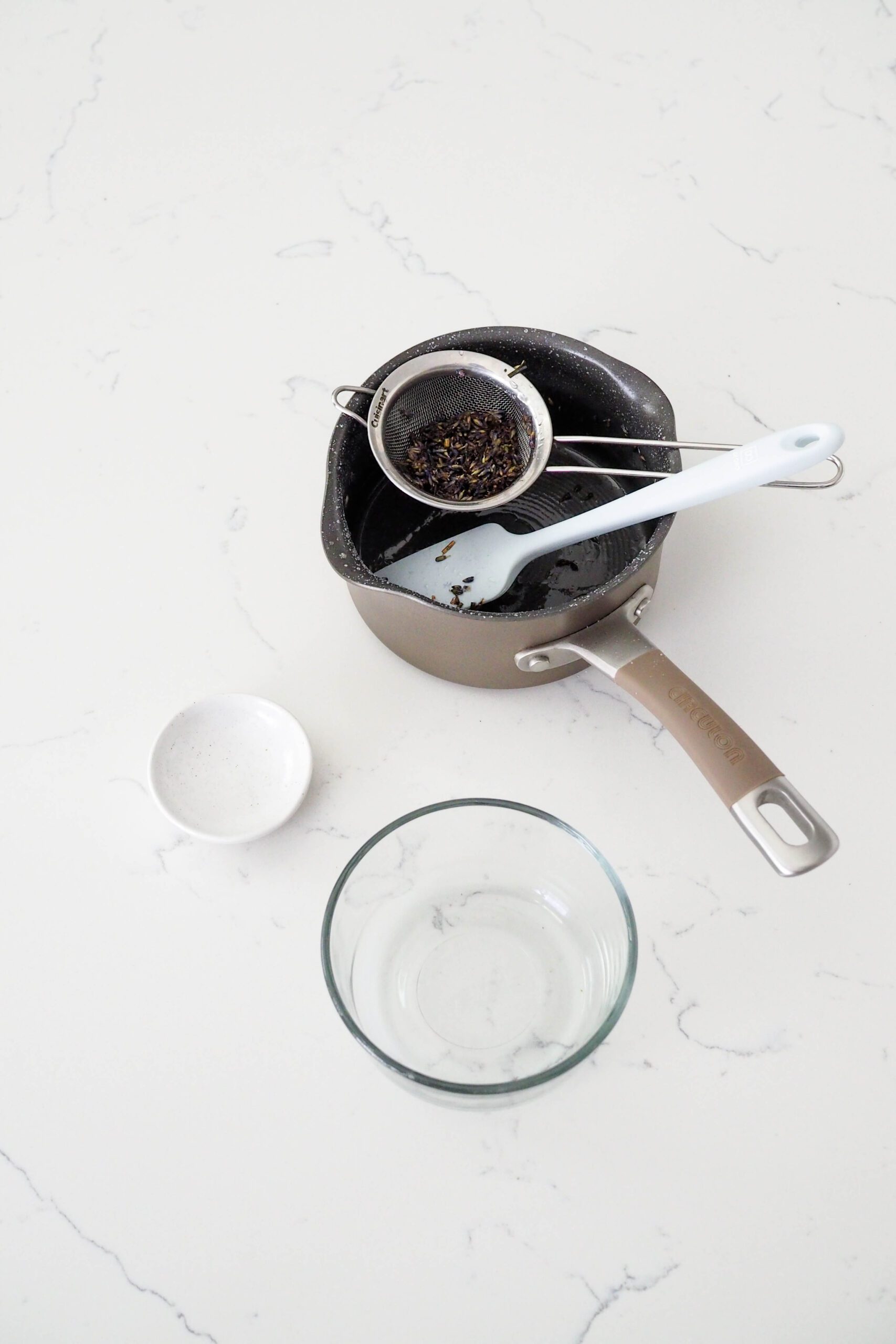 A saucepan, spatula, strainer, and two bowls on a white counter.