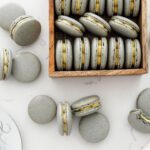 A wooden box of grey macarons is surrounded by more macarons.