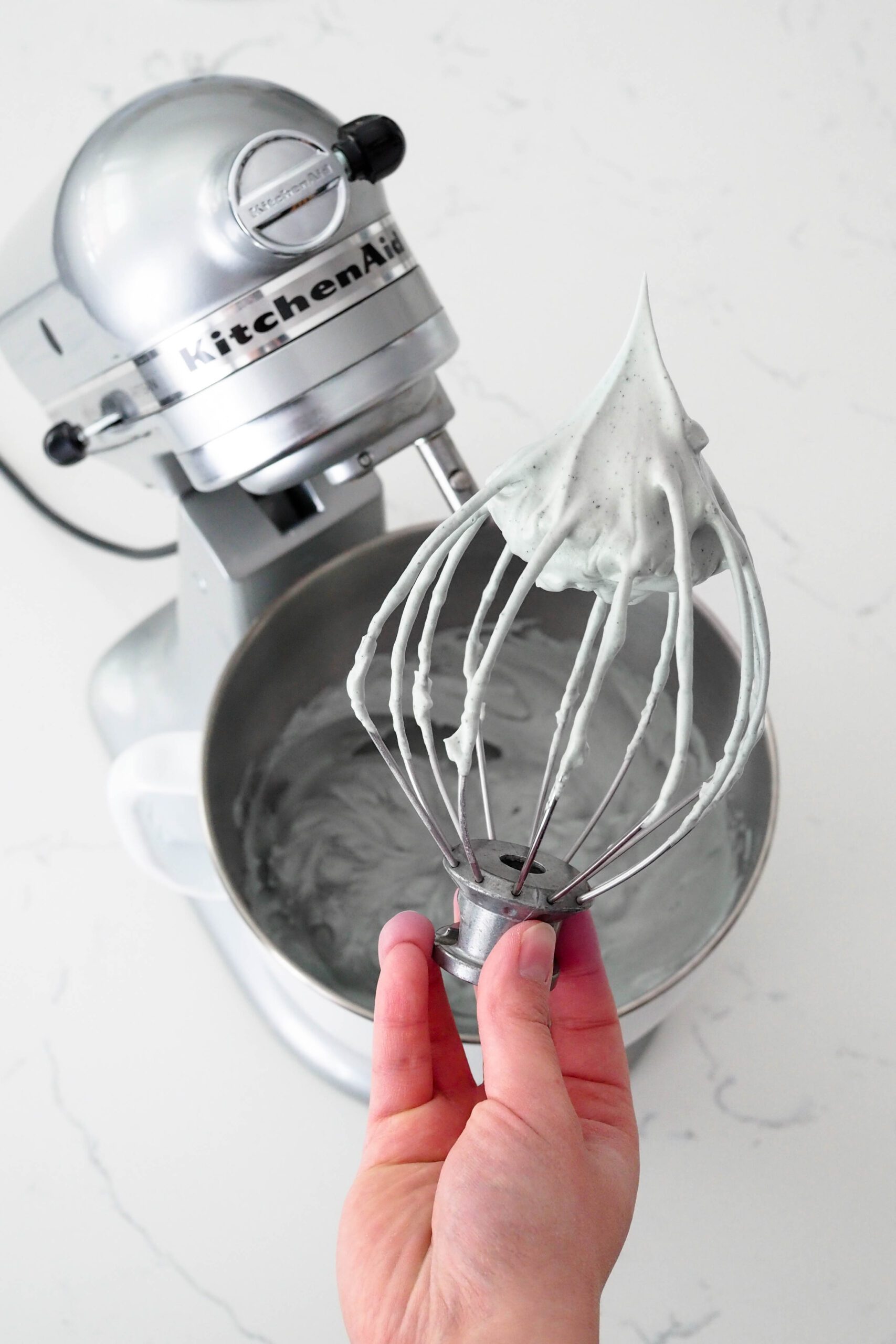 A hand holds up a whisk with grey stiff peaked meringue.