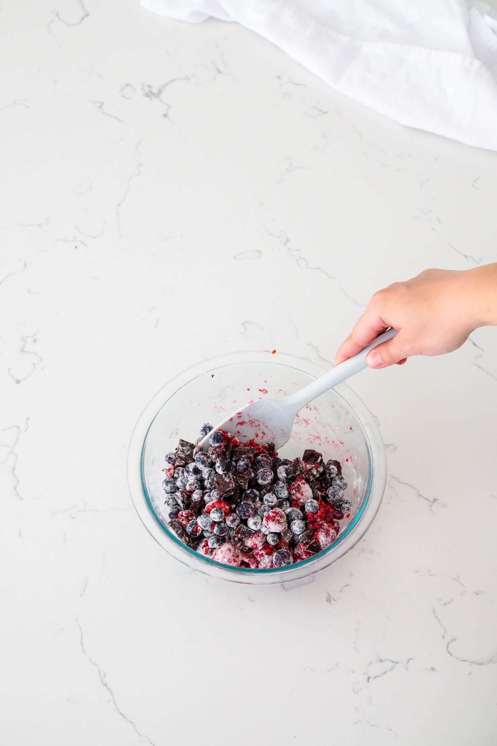 A hand mixes a glass bowl of berries, chocolate, and flour with a spatula.