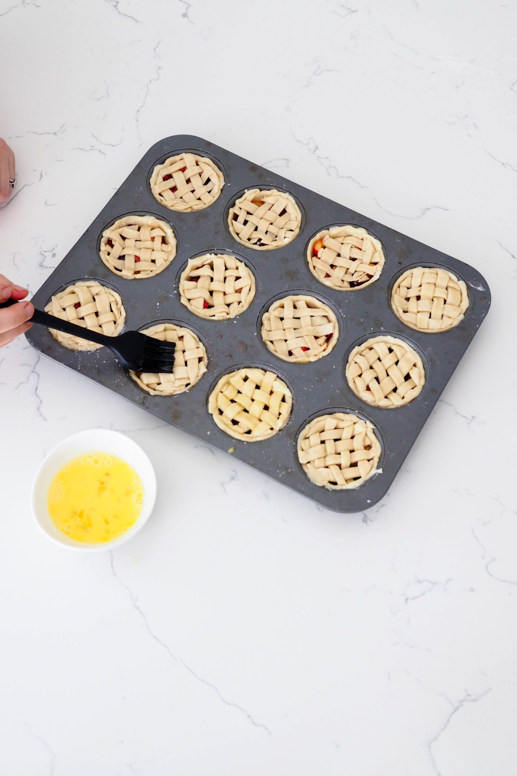A hand uses a silicone pastry brush to brush egg wash onto mini latticed pies.