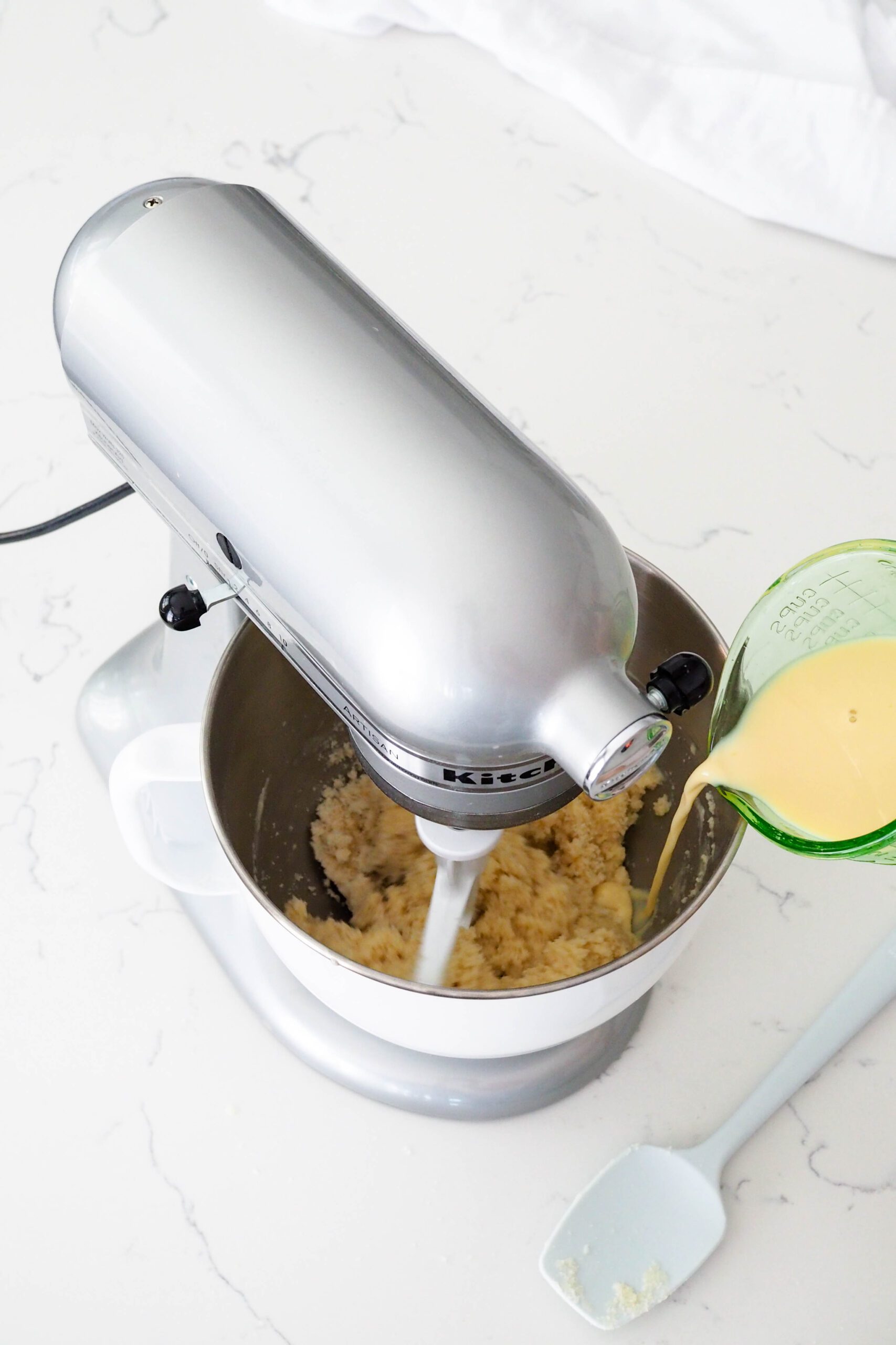 A spouted measuring cup pours wet ingredients into the bowl of a stand mixer.