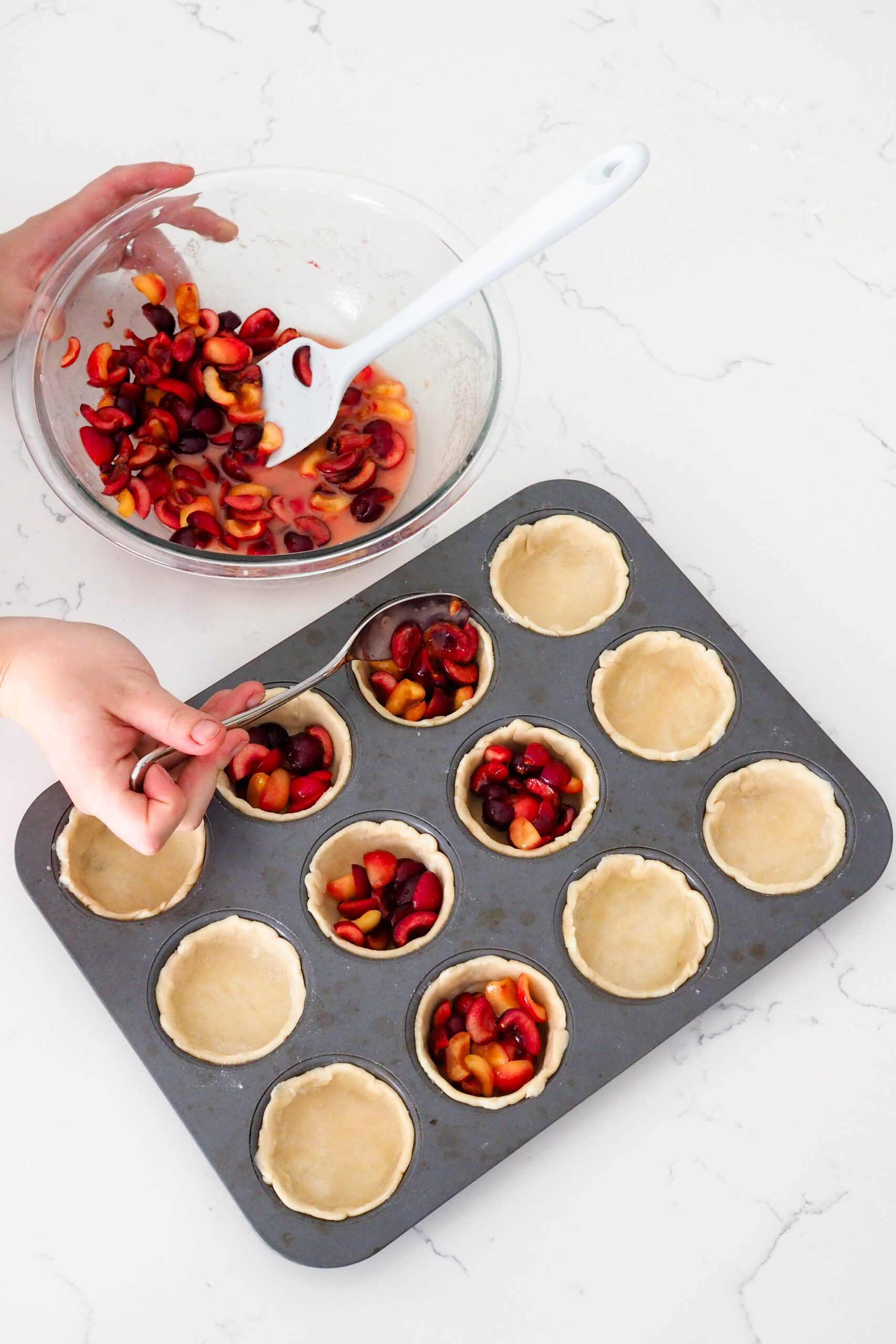 A hand uses a tablespoon to spoon cherries into mini pies.