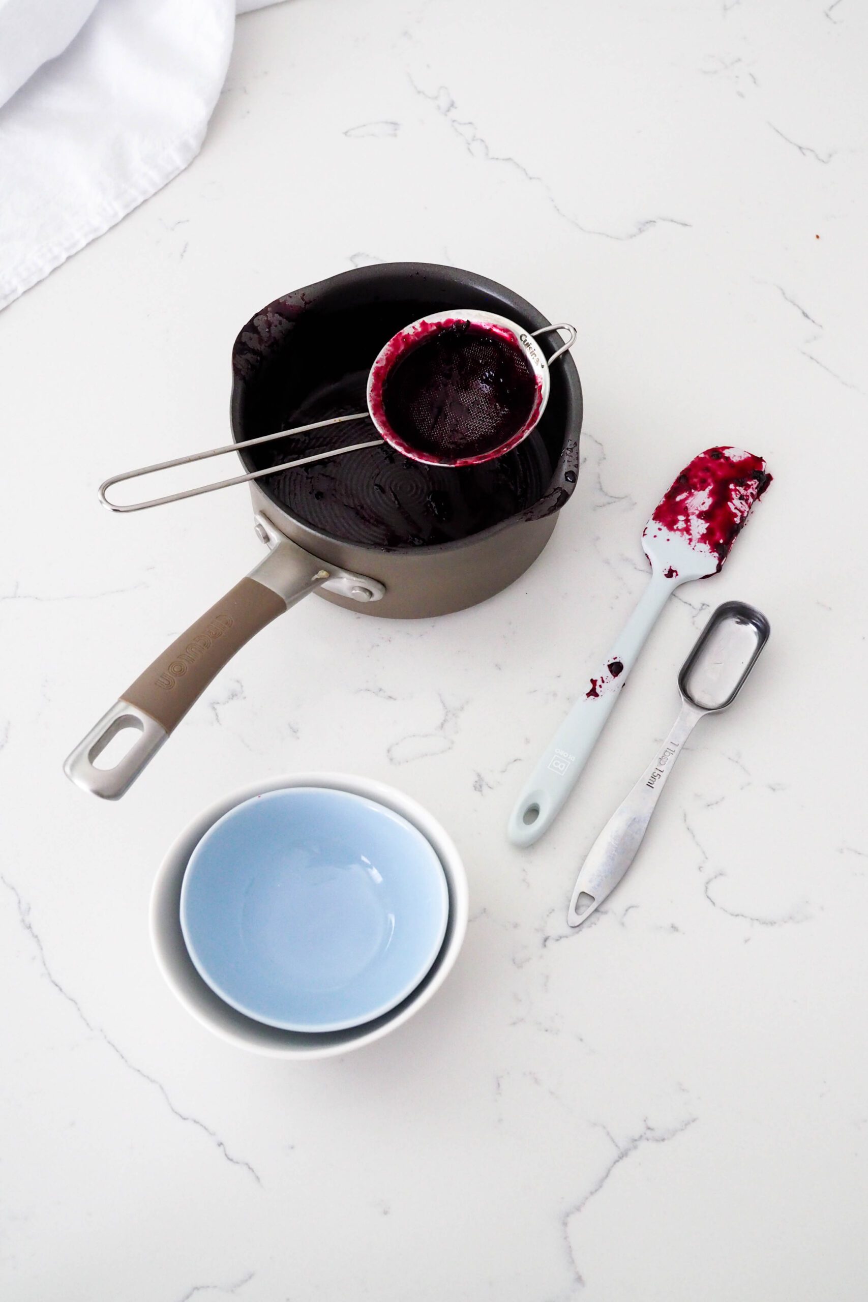 A small pot, strainer, spatula, measuring spoon, and two small bowls on a counter with remnants of blueberry syrup.