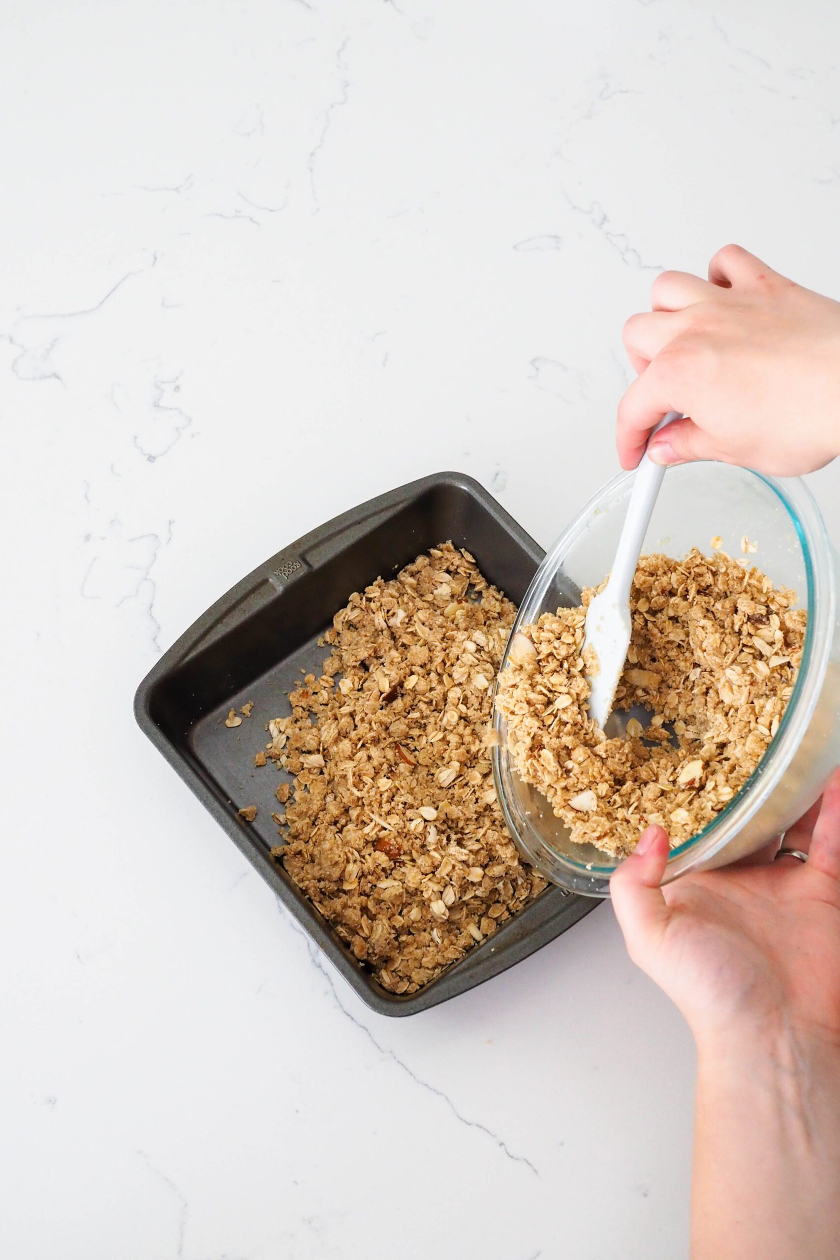 Two hands pour gluten-free crumble crust into a square pan.