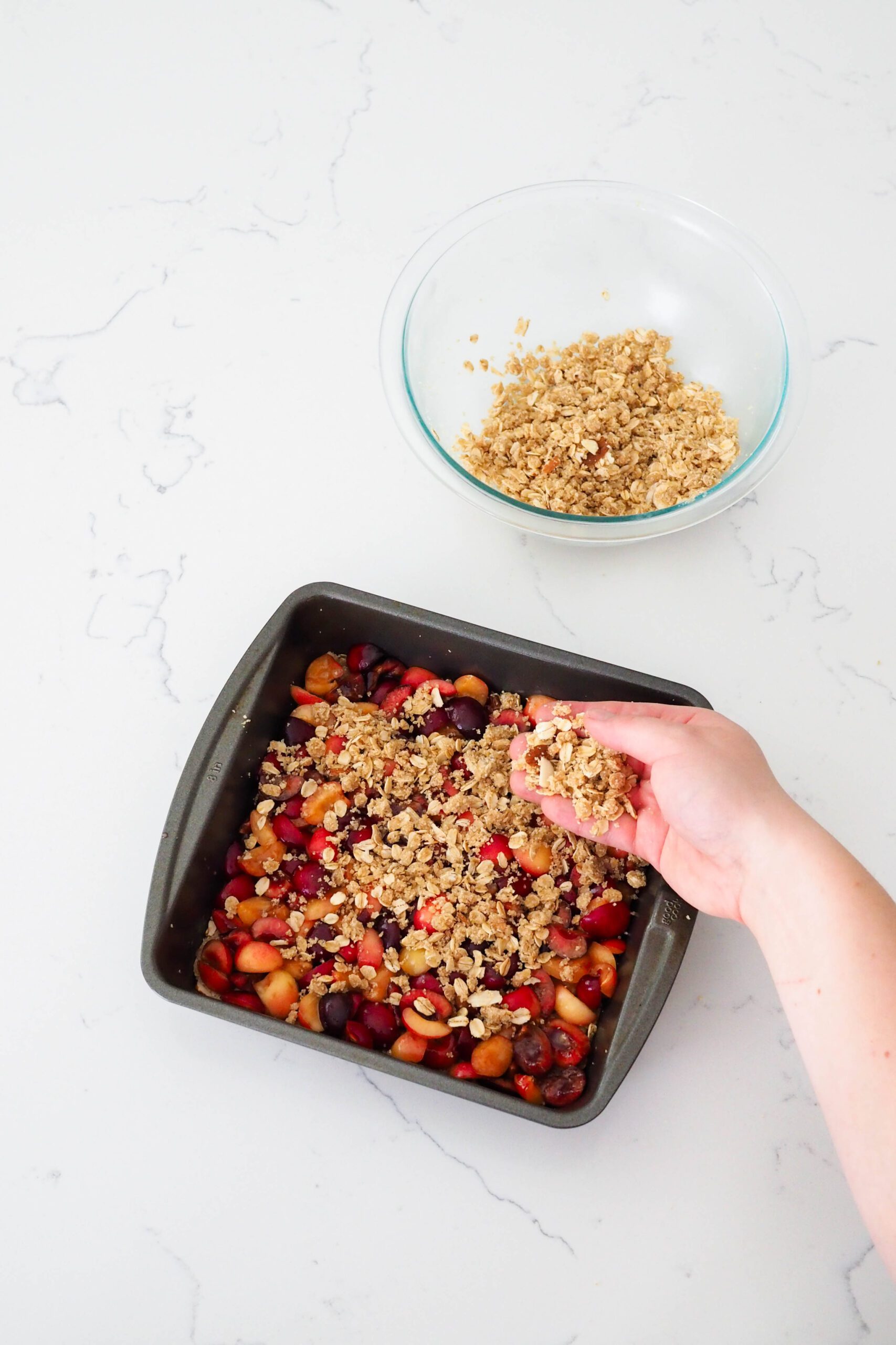 A hand sprinkles gluten-free crumble on top of a cherry layer in a square pan.