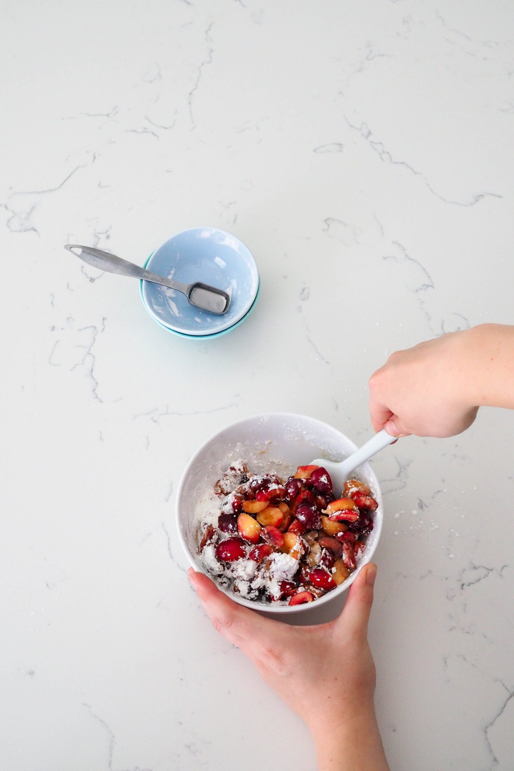 Two hands stir together cherries, sugar, almond extract, and cornstarch in a glass bowl.