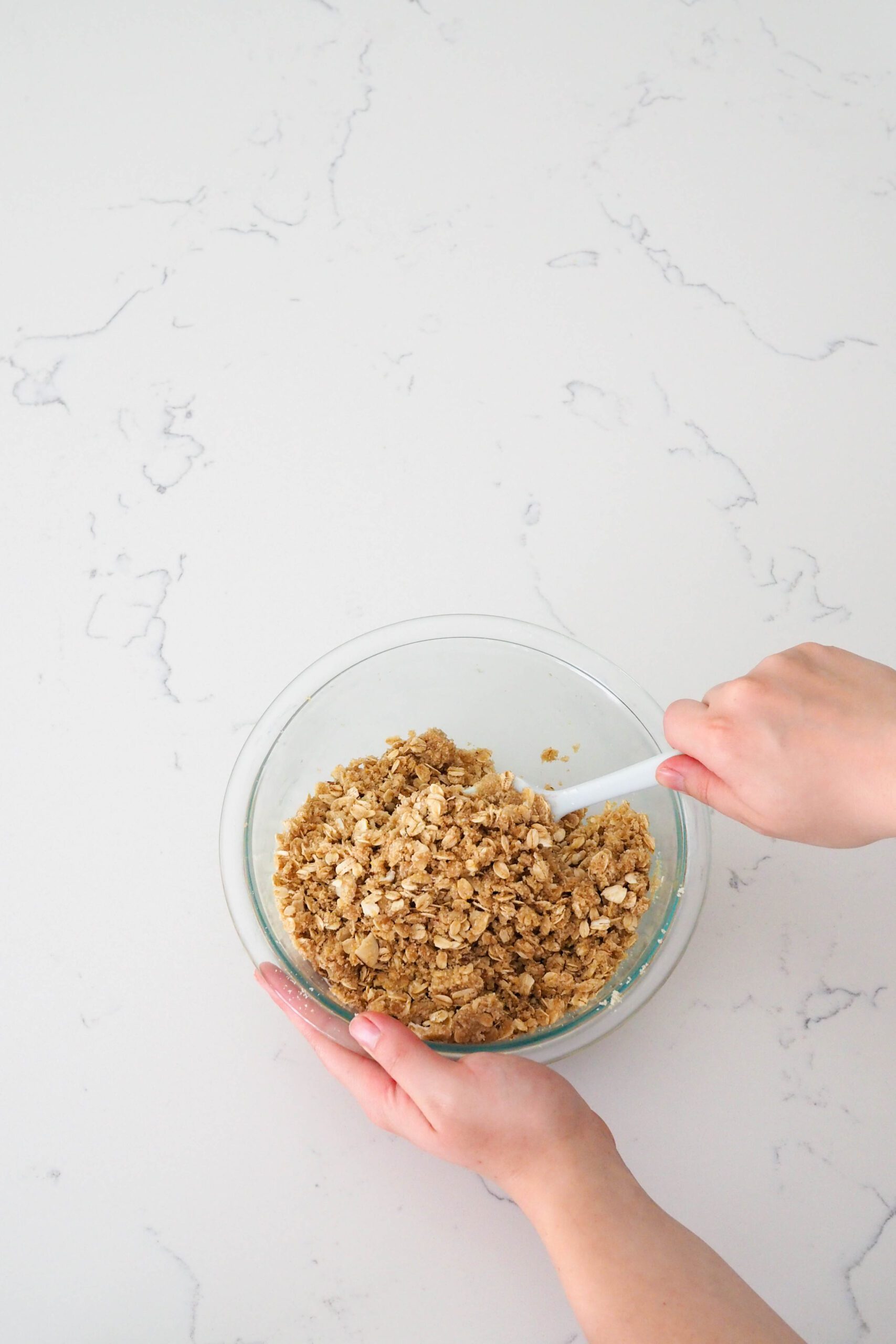 Two hands stir together oats and dry ingredients in a glass bowl.