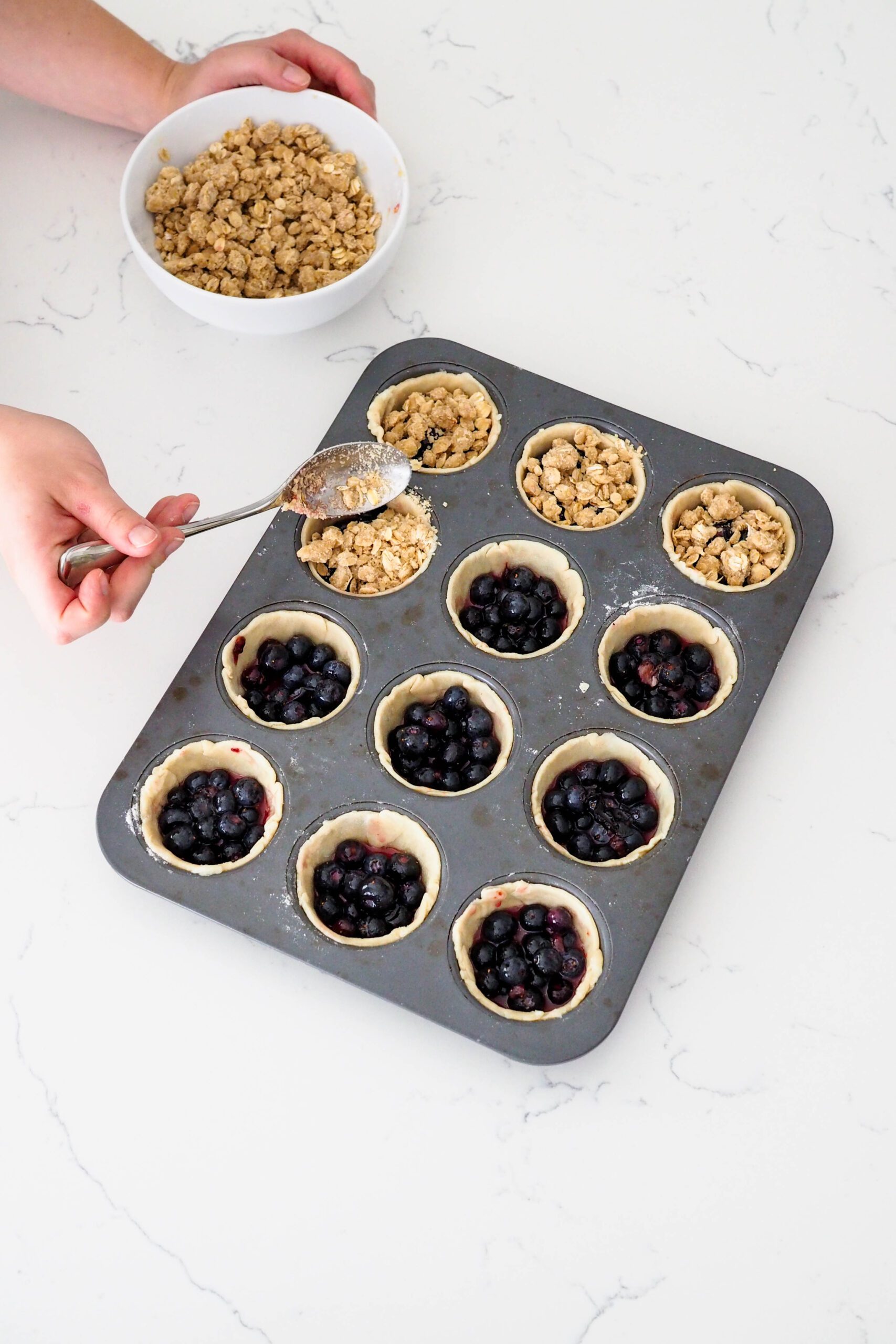 A hand spoons crumble onto mini blueberry pie shells.