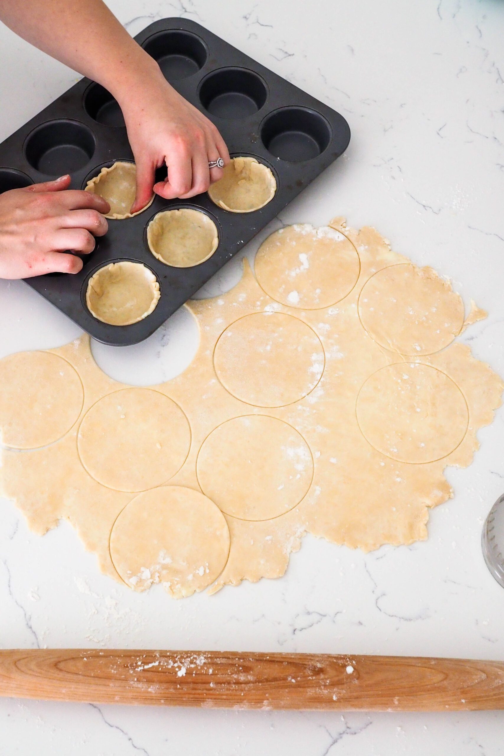 Two hands adjust the edges of a pie dough round in a muffin pan hole.