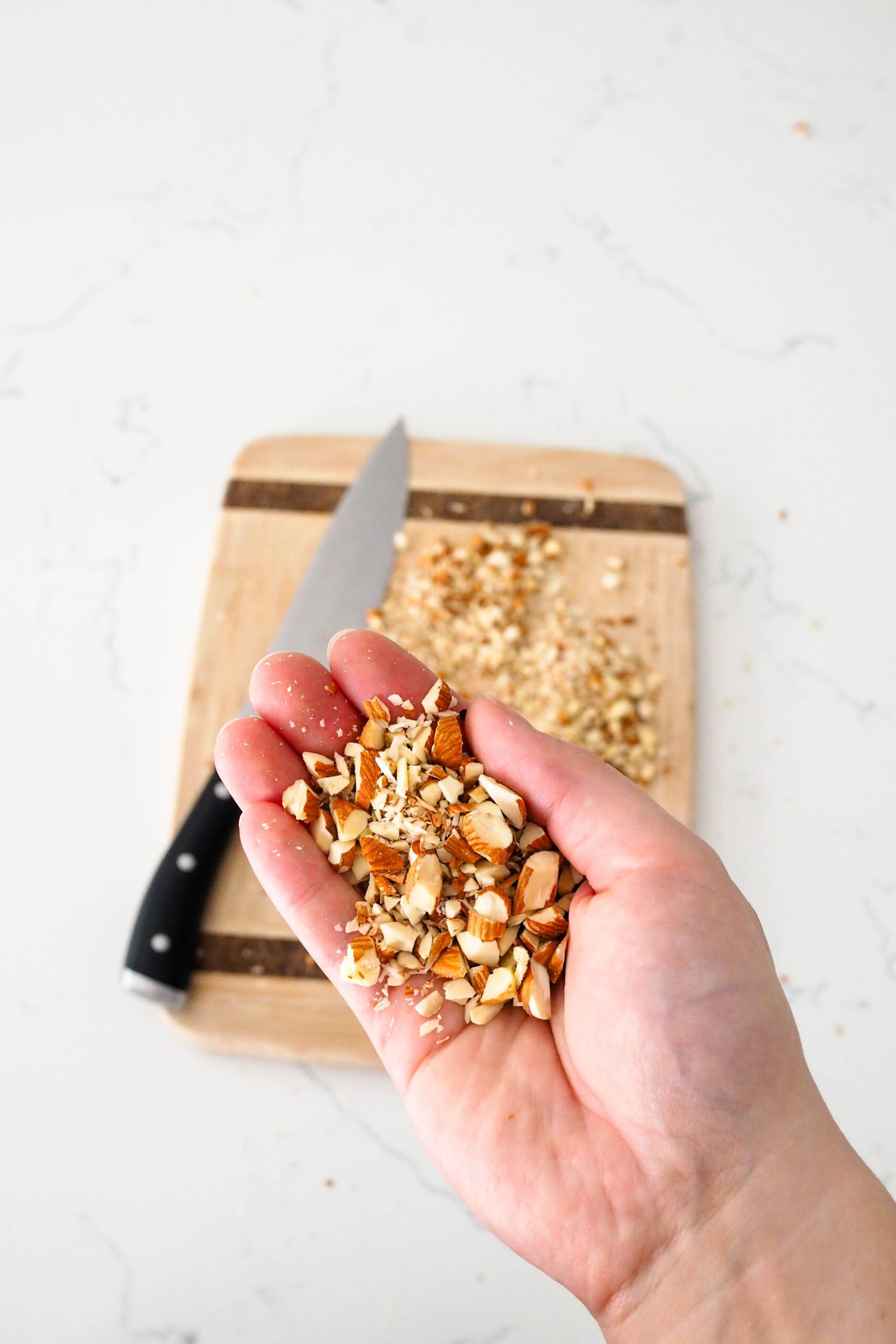 A hand holds up chopped almonds in front of a cutting board and knife.