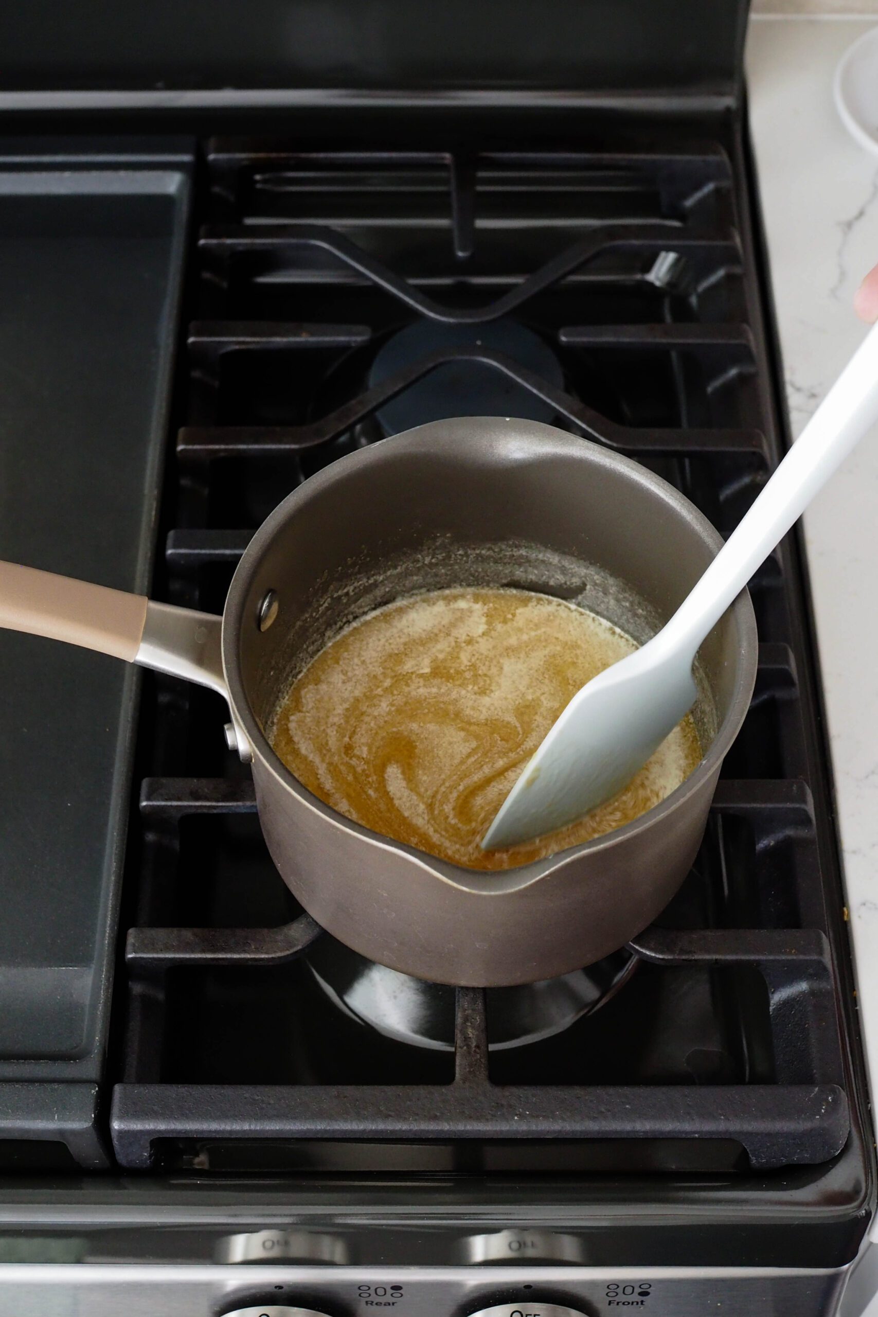 A spatula stirs melted butter, honey, and brown sugar in a pot over the stove.
