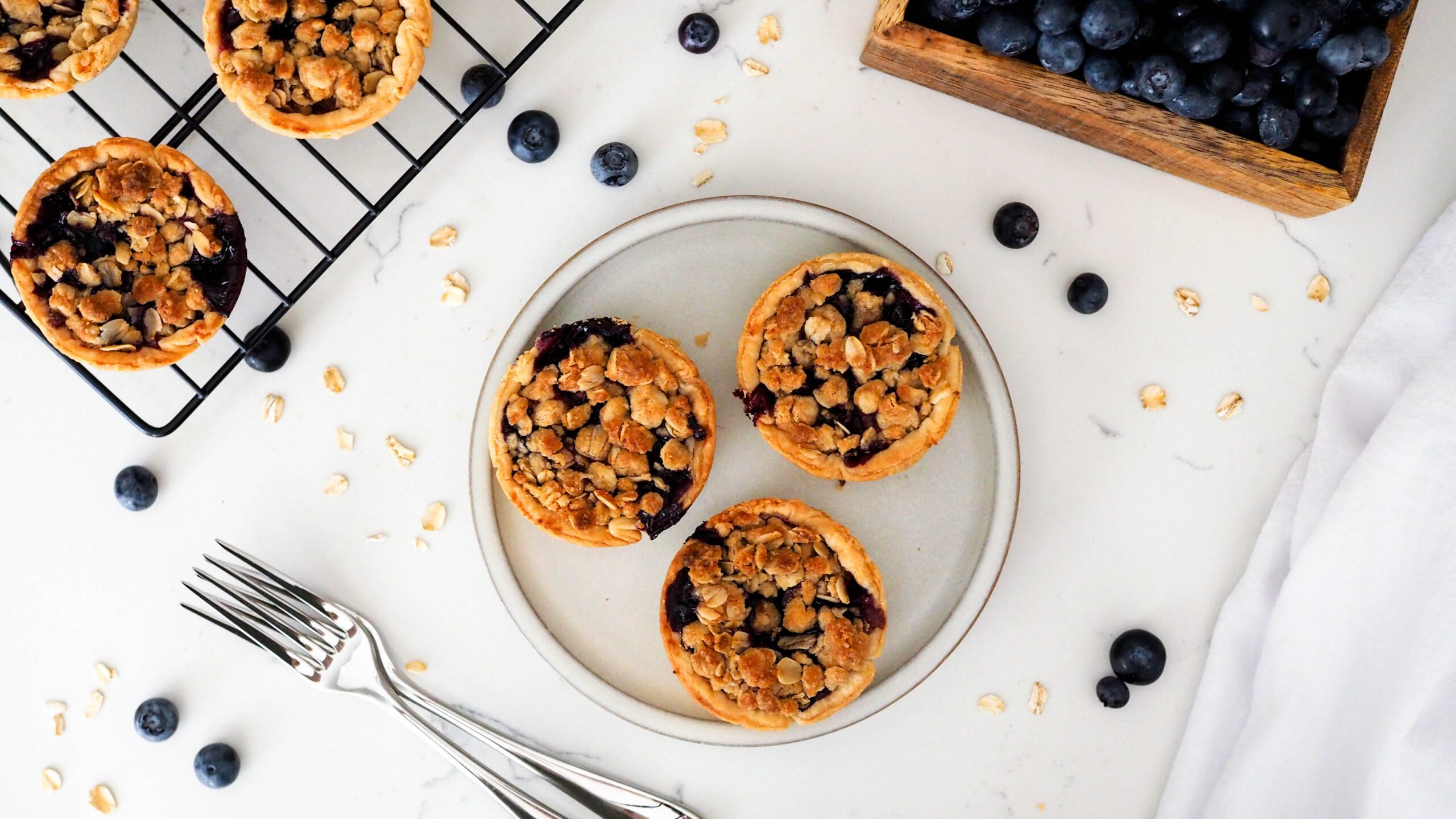 An overhead view of three mini blueberry pies on a small plate with blueberries and oats nearby.