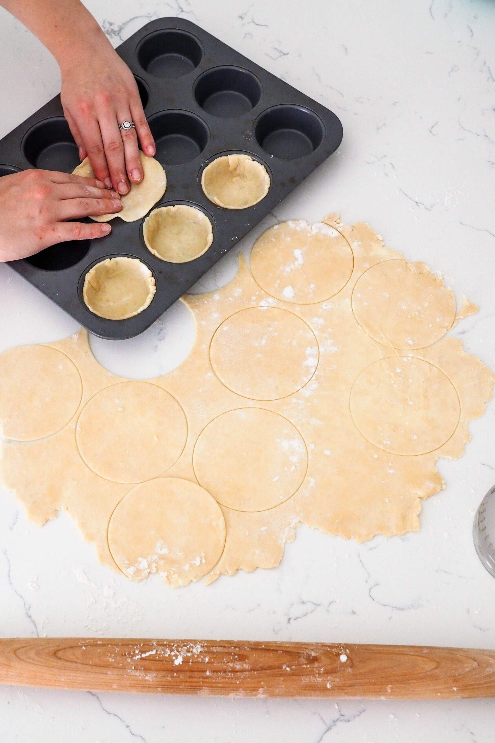 Two hands guide a circle of pie dough into a muffin pan opening.