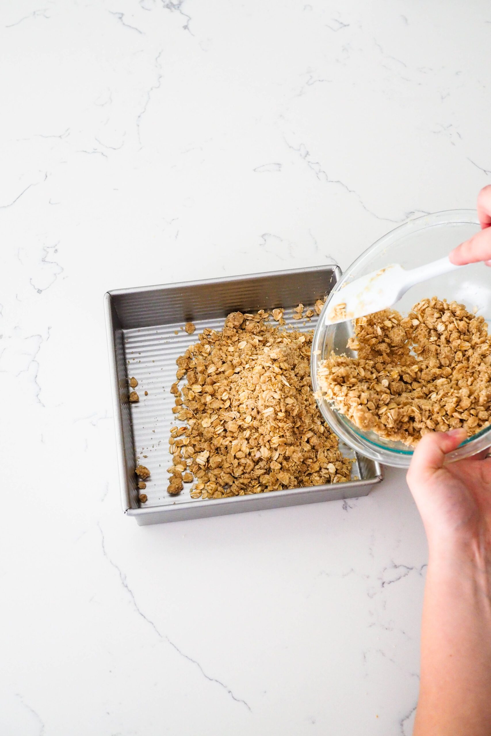 Two hands pour an oat crumble crust into a square pan.