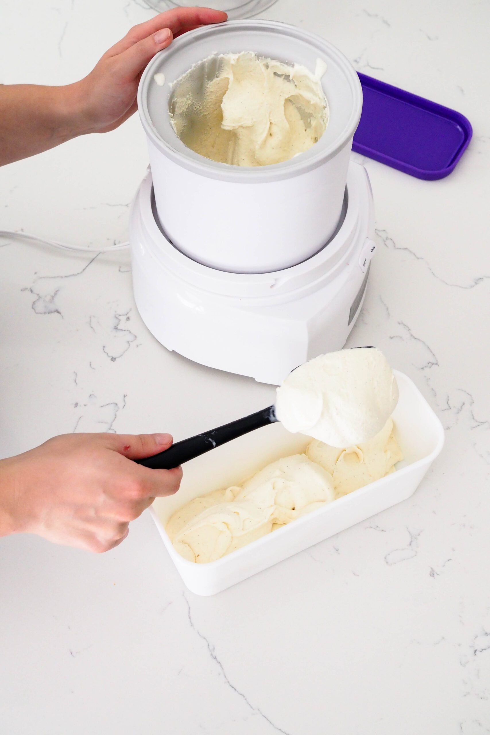 A hand scoops ice cream out of an ice cream maker canister and into a rectangular container.
