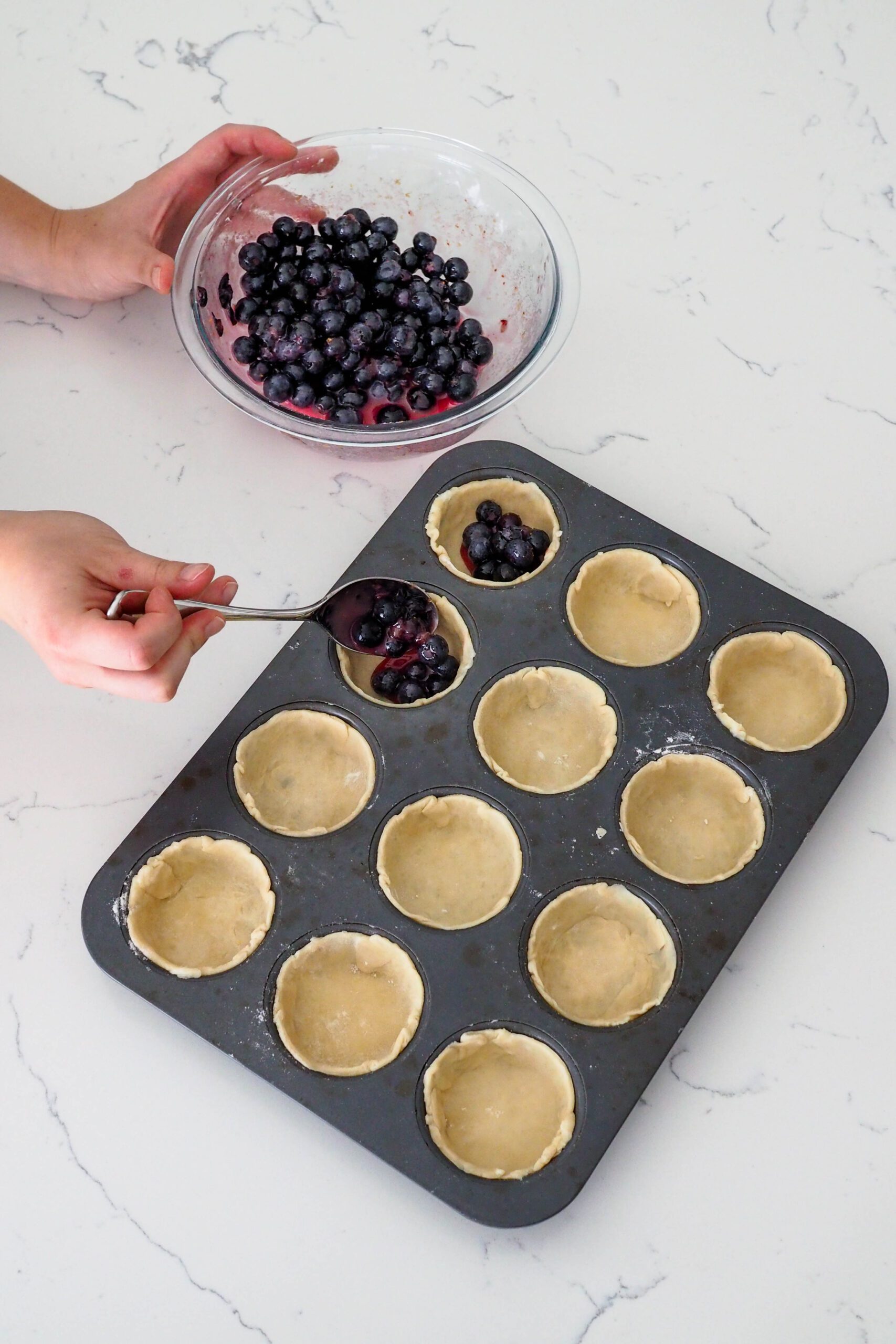 A hand spoons blueberry pie filling into mini blueberry pie shells.