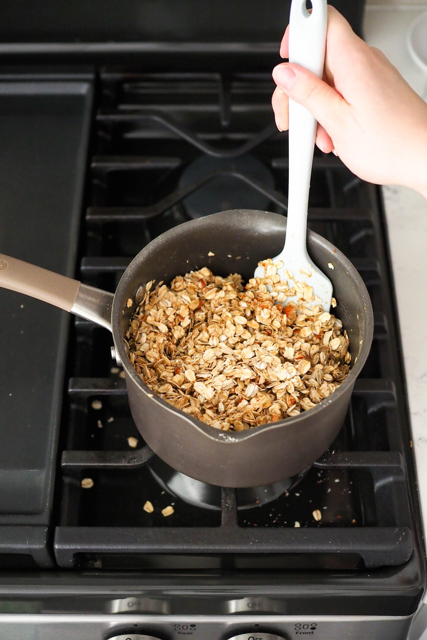 A hand stirs together granola in a pot with a spatula.