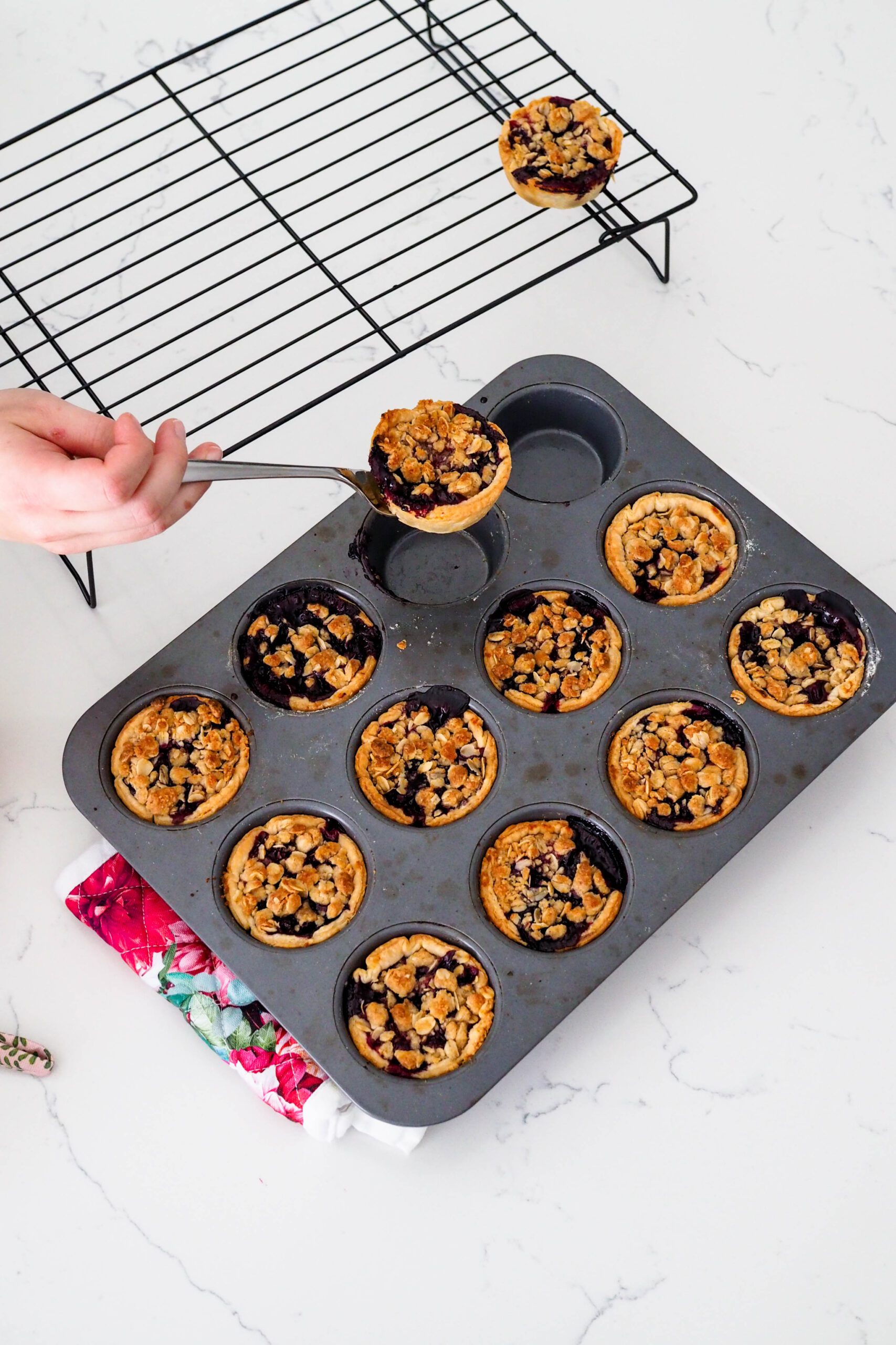 A mini pie is removed from a muffin pan with a fork and placed on a wire rack.