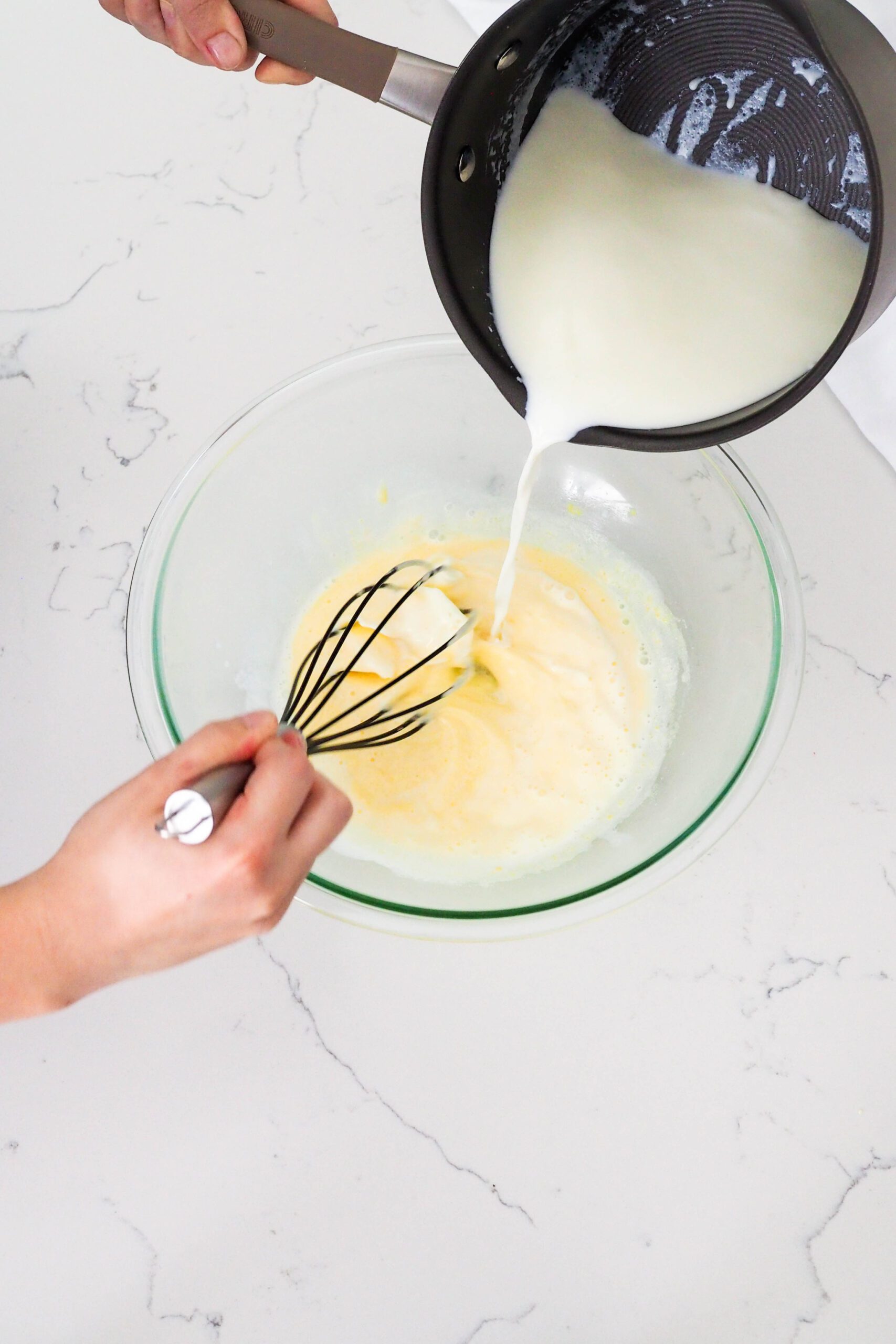 Hot milk and cream from a pot is whisked into egg yolks at the ribbon stage.