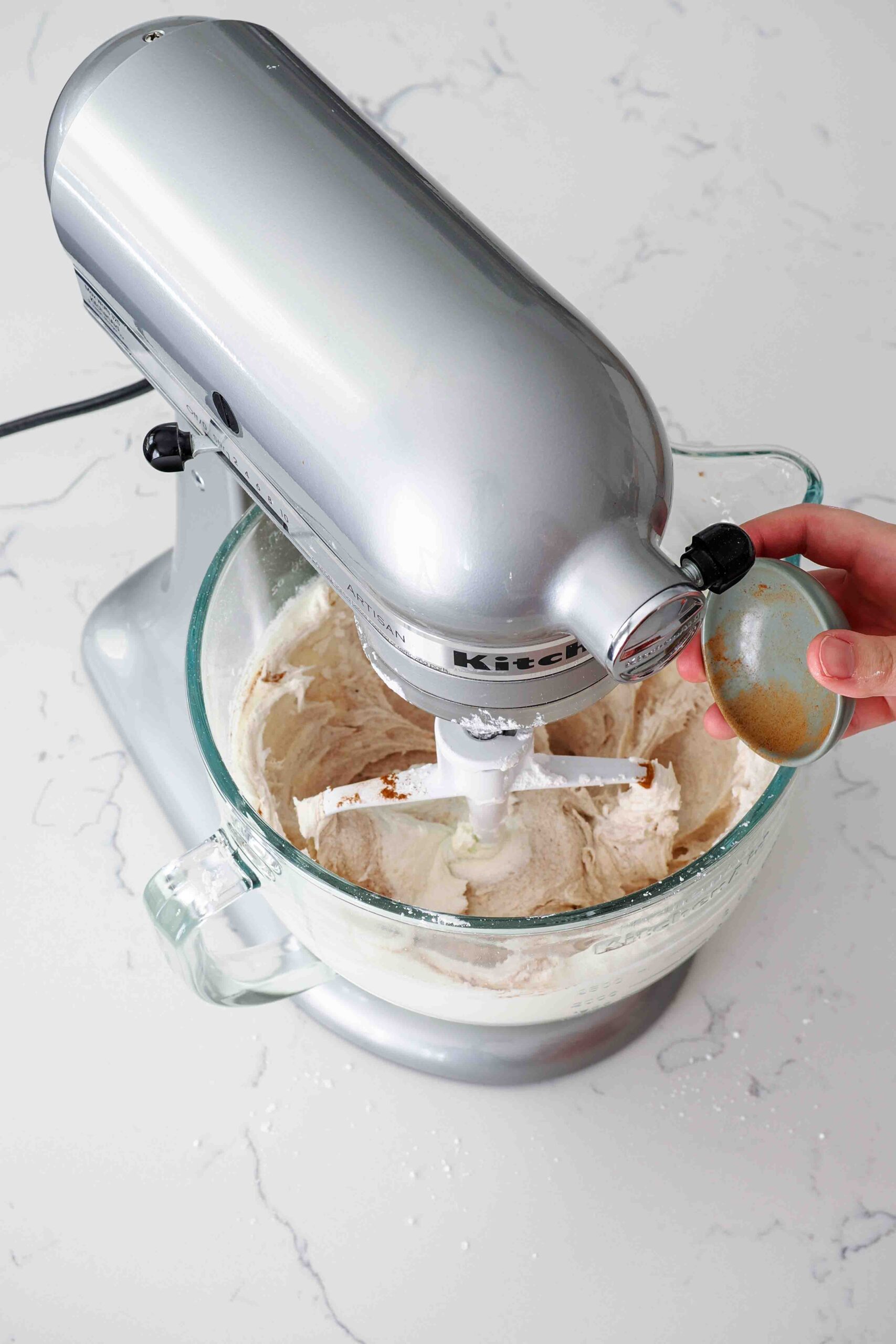 A hand adds ground cinnamon to buttercream in a stand mixer bowl.