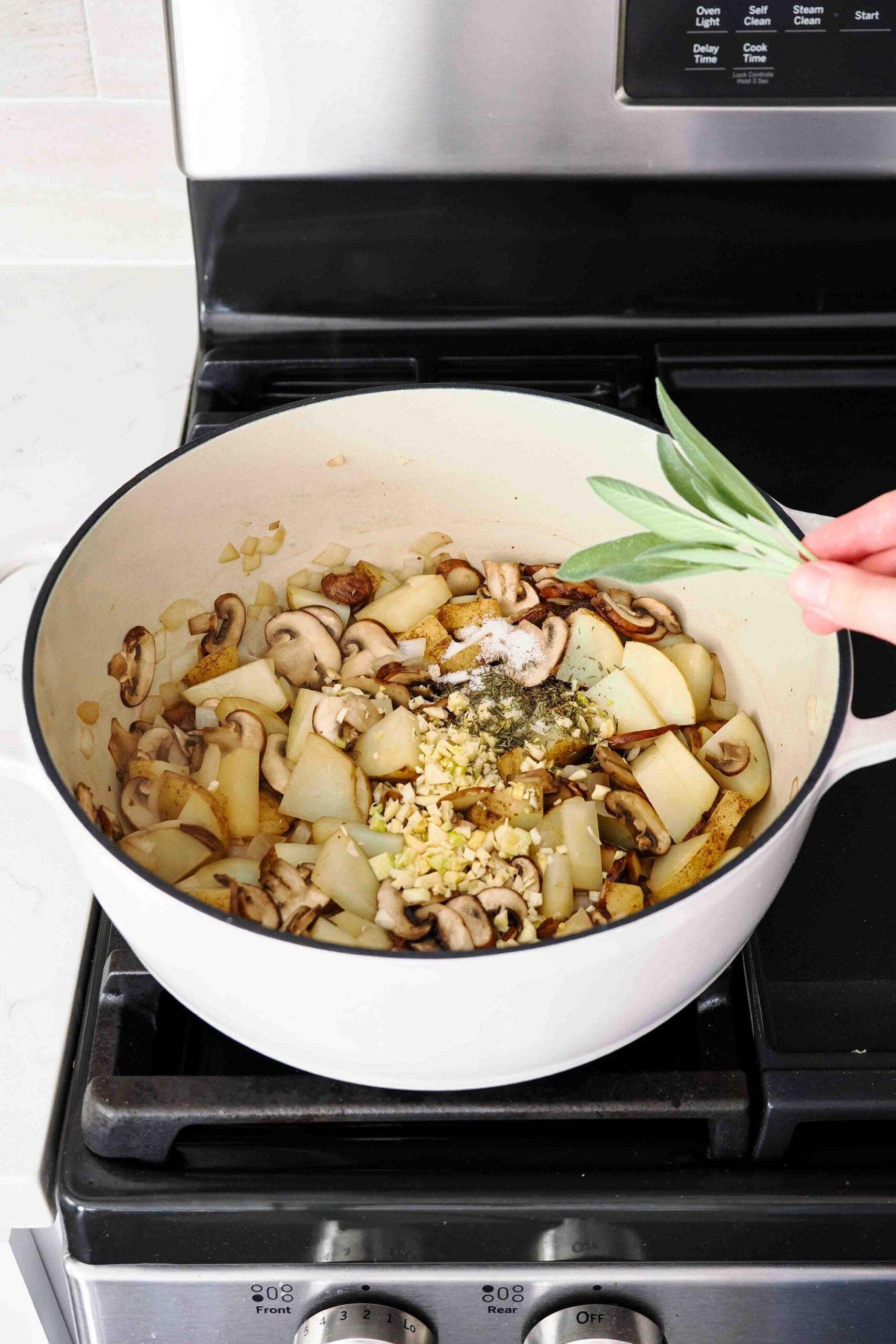 A hand adds a bunch of fresh sage to cooked vegetables in a Dutch oven.