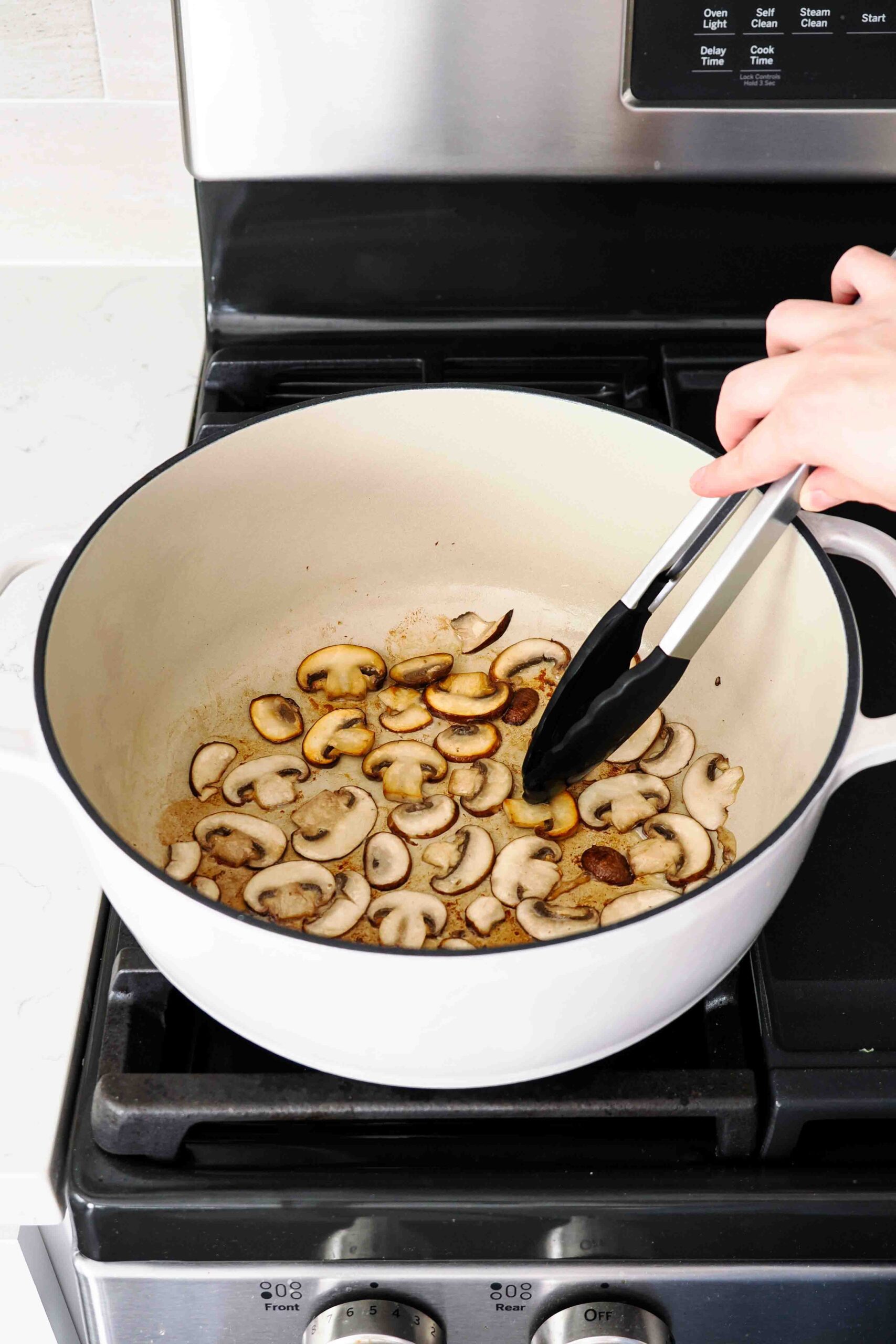 A hand uses tongs to flip over sliced mushrooms in a Dutch oven.