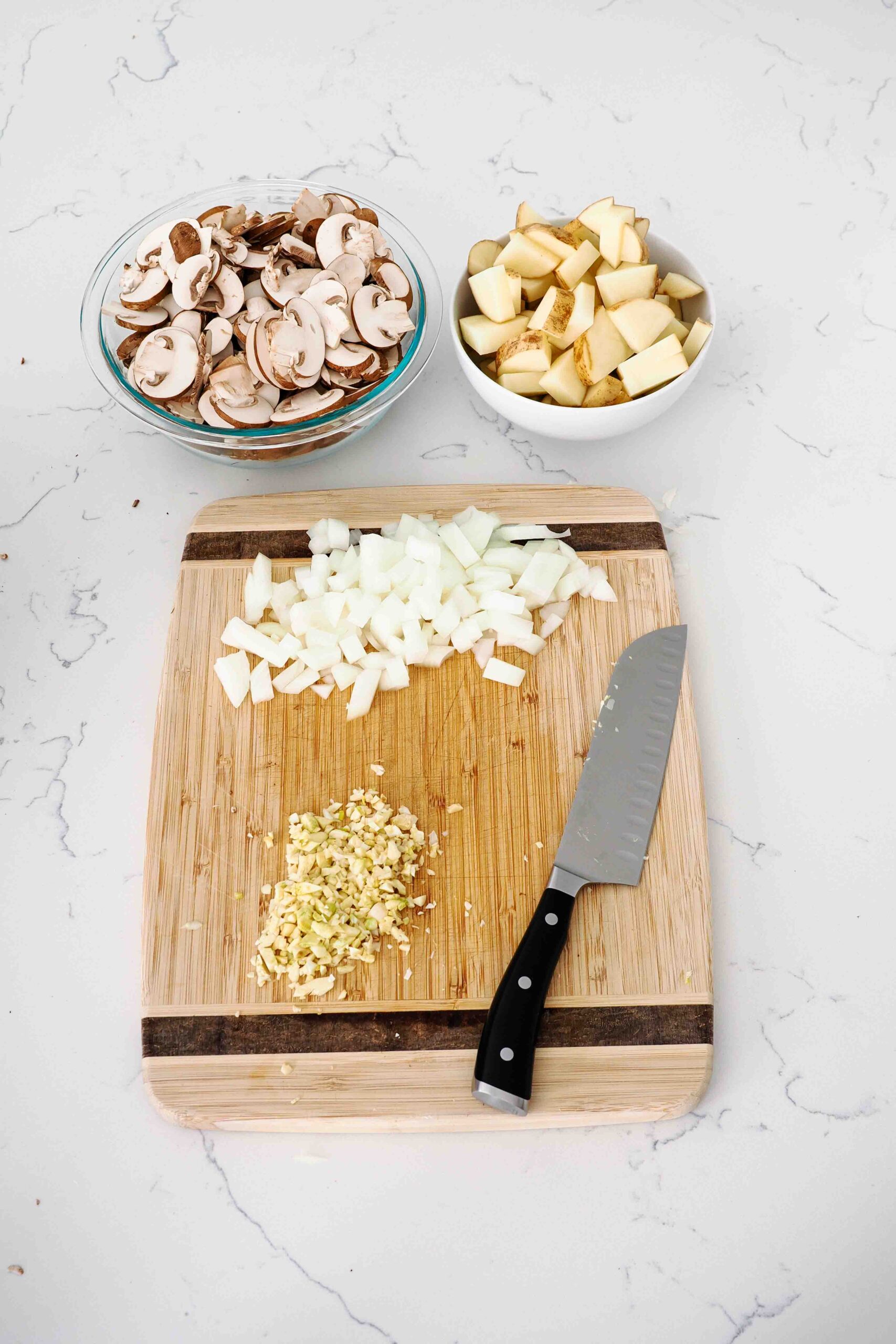 A cutting board and knife with chopped mushrooms, potatoes, onion, and garlic.