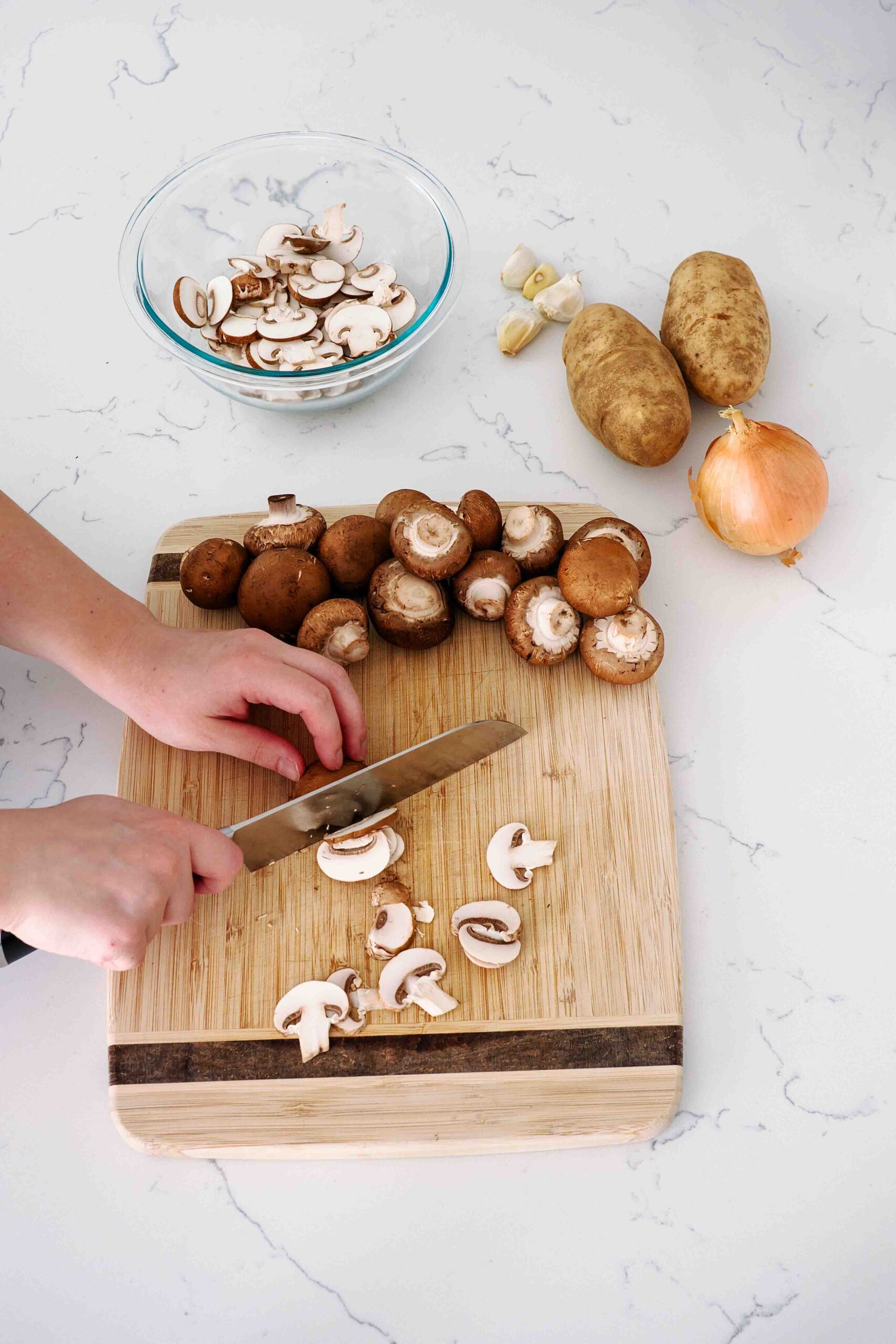 Two hands thinly slice mushrooms with a knife on a cutting board.