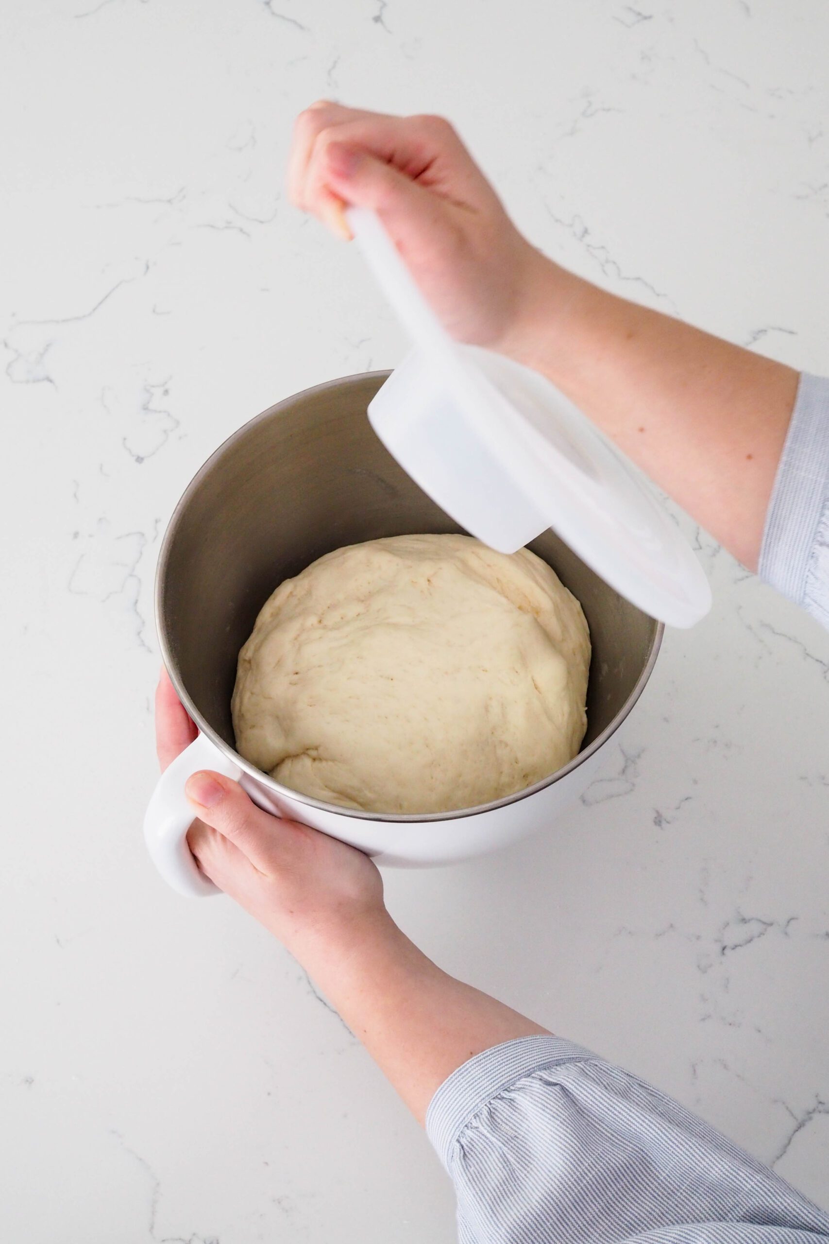 A hand removes the lid off of a mixer bowl with enriched dough that has doubled in size.