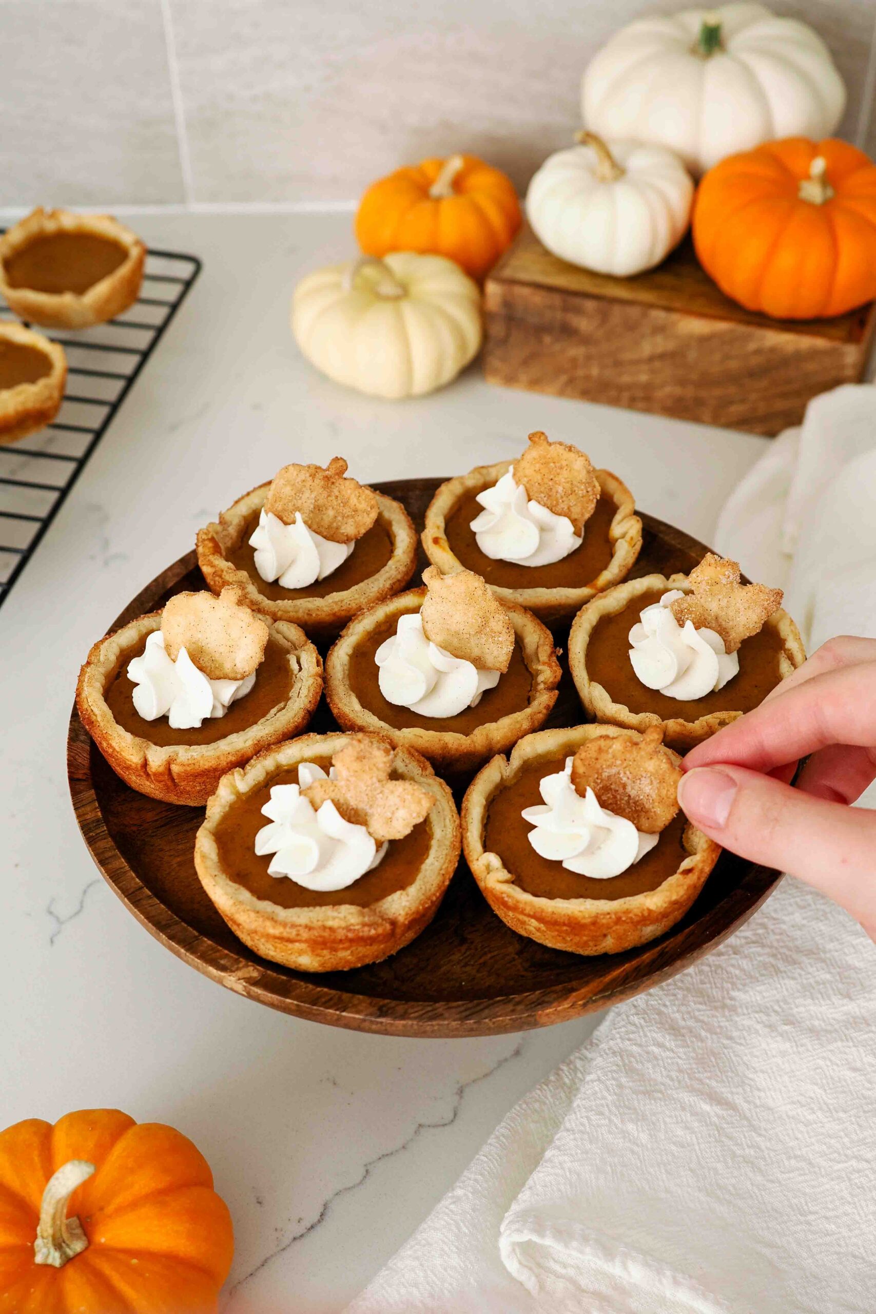 A white hand adds decoration to a mini pumpkin pie with whipped cream on a small cake stand.