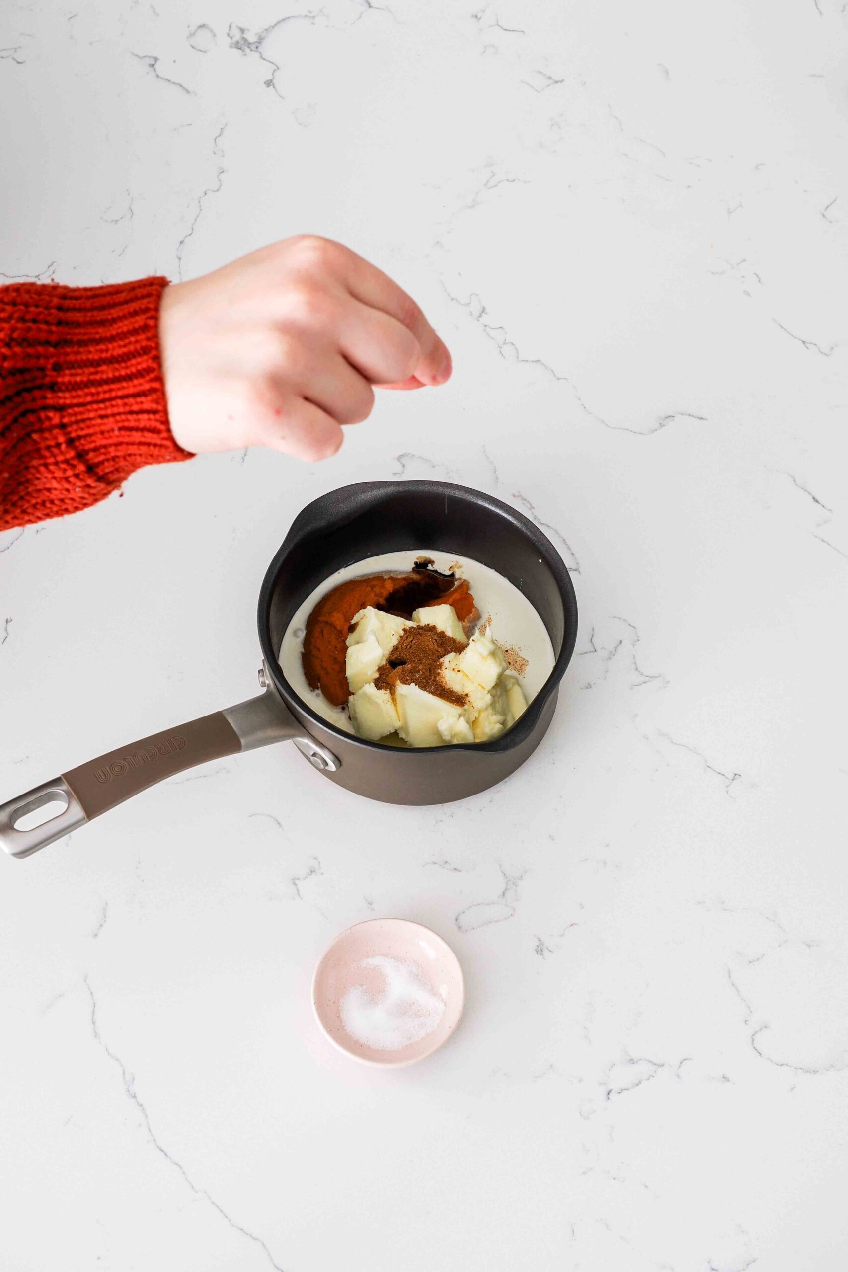A hand adds a pinch of salt to a small saucepan with pumpkin, butter, and other ingredients.