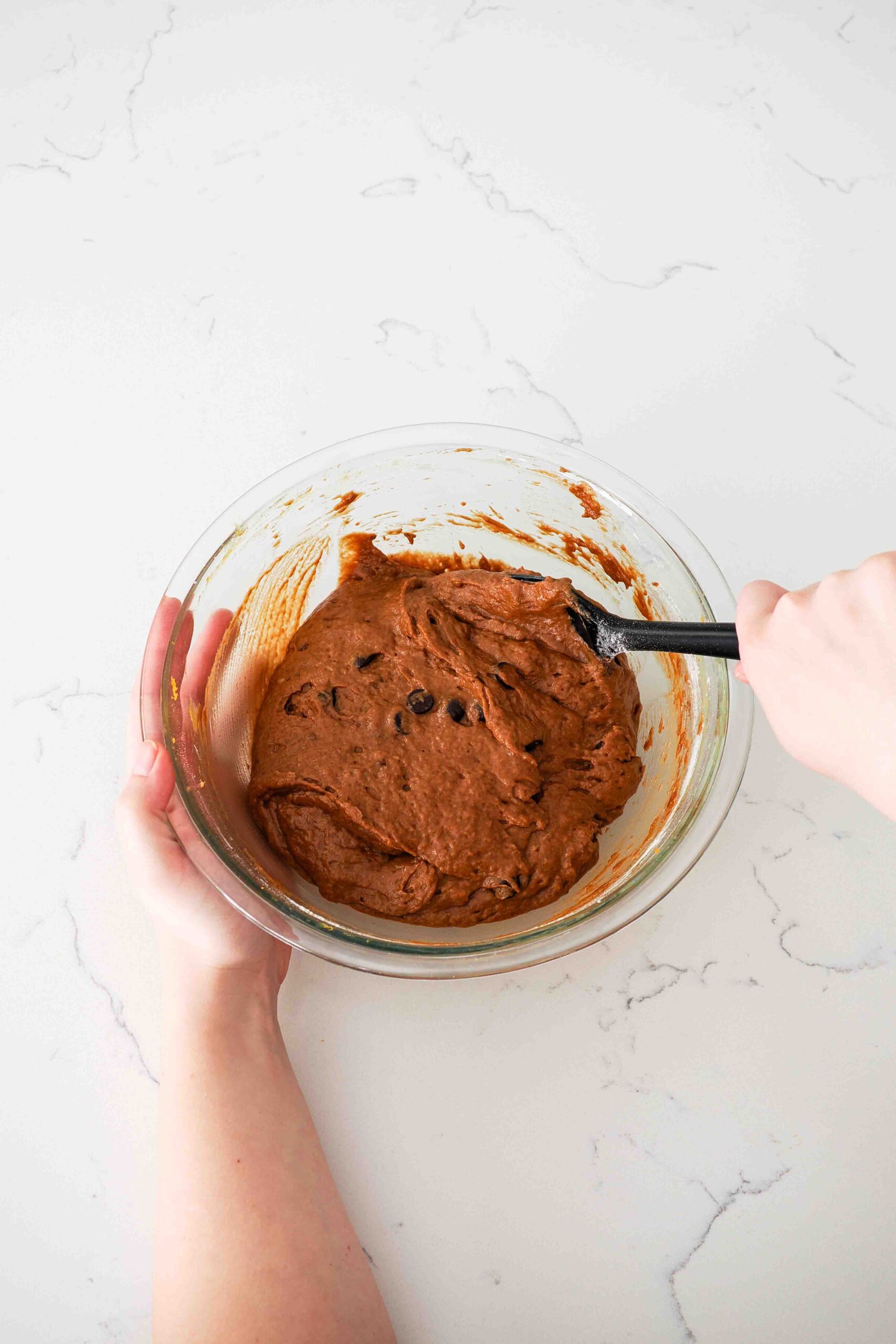 Two hands use a spatula to stir together chocolate pumpkin muffin batter.