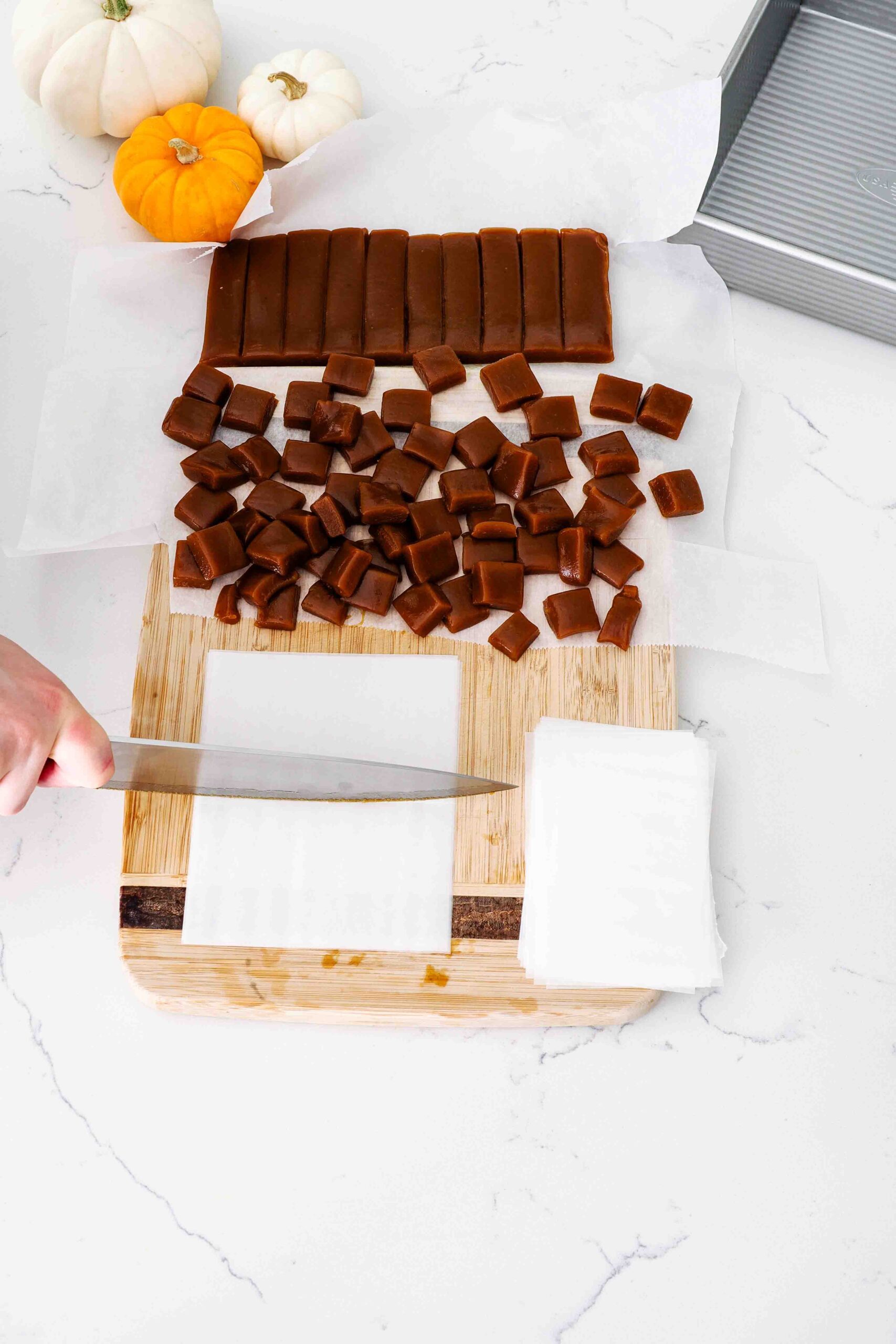 A knife cuts pre-cut wax paper pieces in half on a cutting board with caramels.