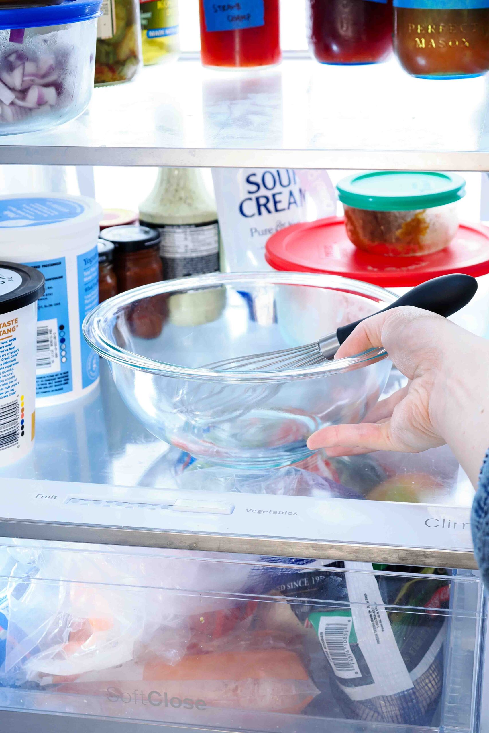 A hand places a medium glass bowl and whisk in the fridge.