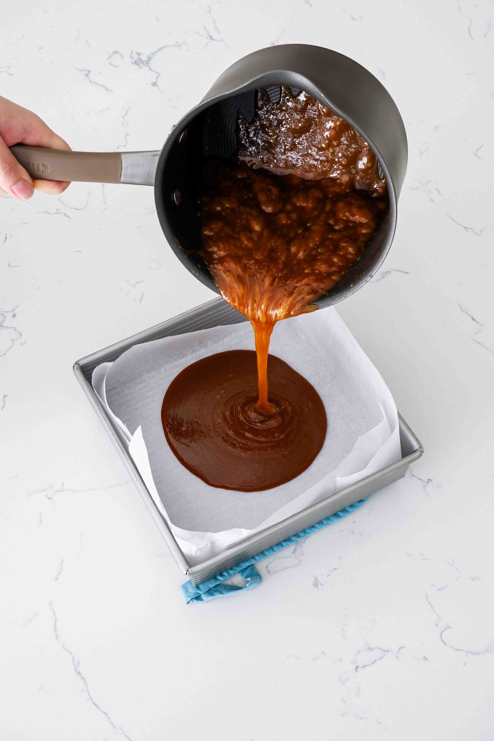 A hand pours pumpkin spice caramel into a parchment-lined pan.