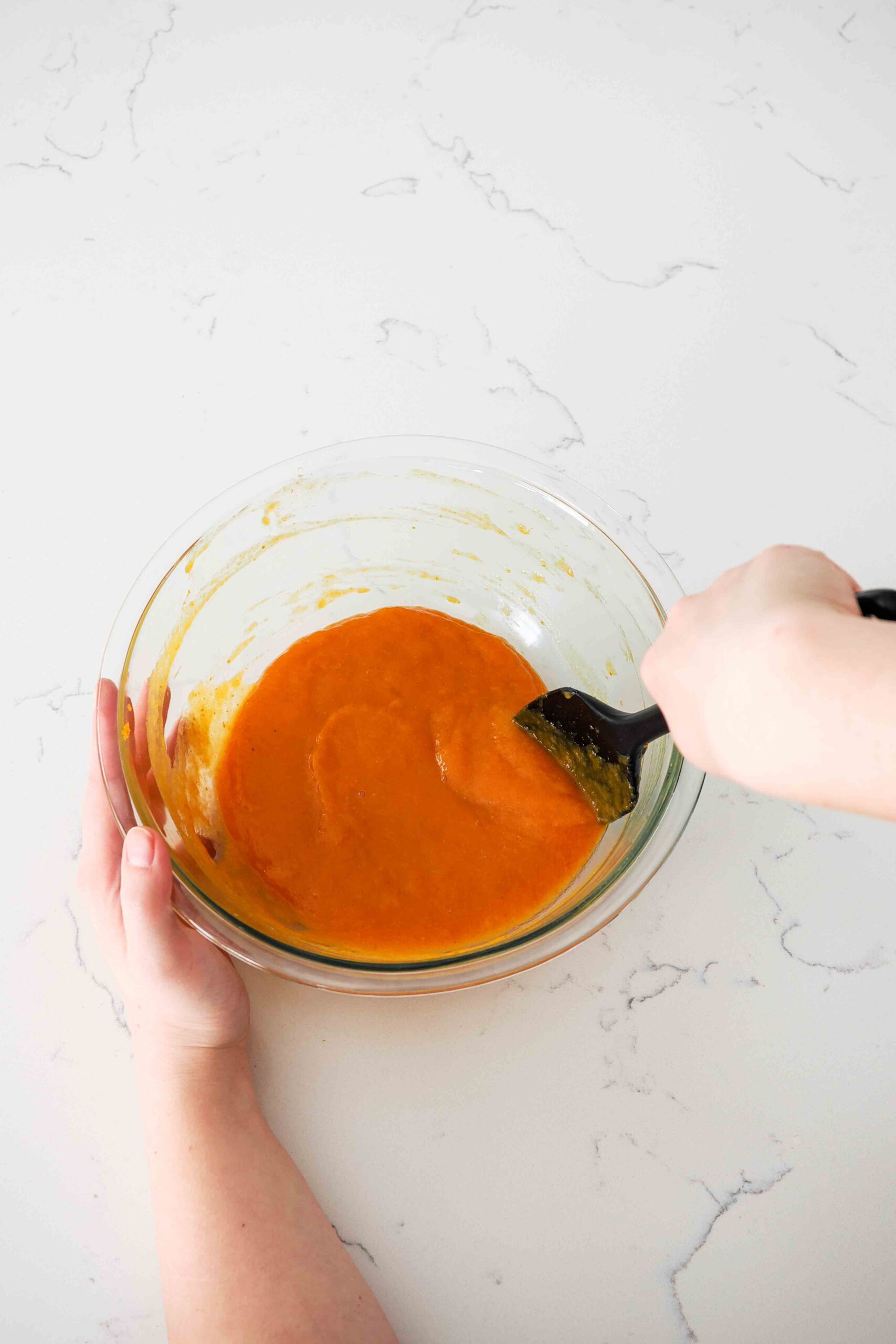 A hand uses a spatula to stir together a wet, pumpkin-colored batter.